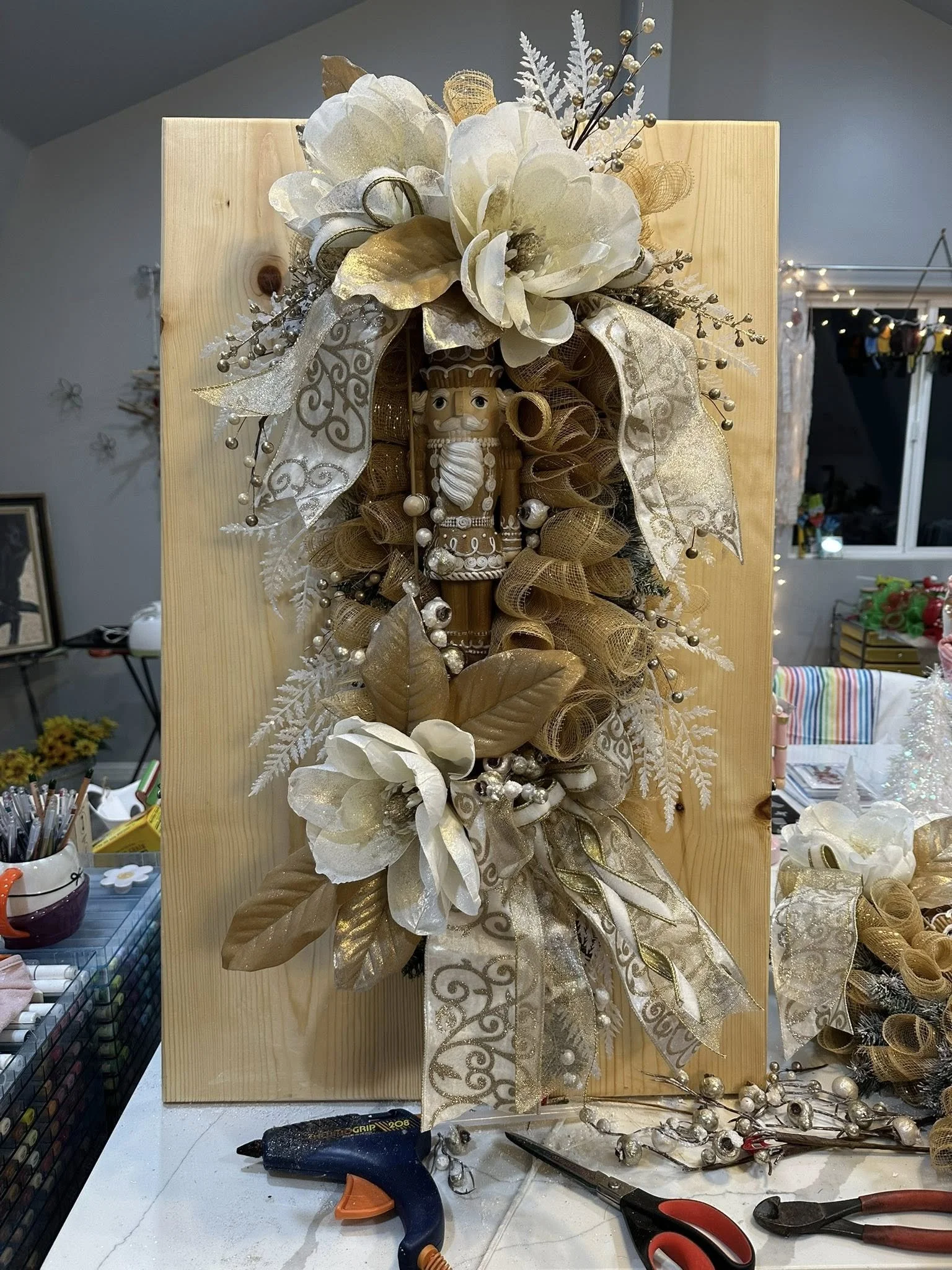 Decorative holiday arrangement with white flowers, gold leaves, ribbons, and a nutcracker figurine on a wooden board.