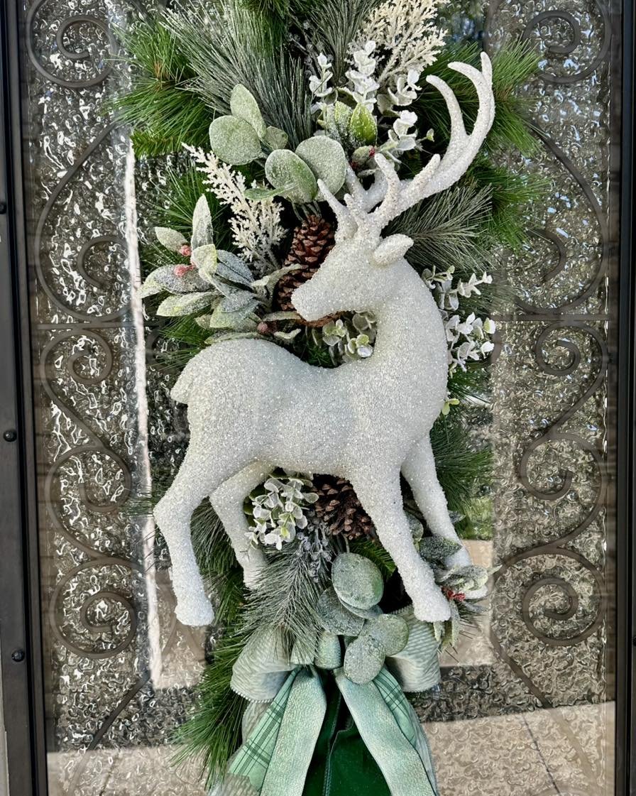 Decorative holiday wreath with a glittery white reindeer in the center, surrounded by pine branches, pinecones, and silver and green ornaments, adorned with a green plaid ribbon.