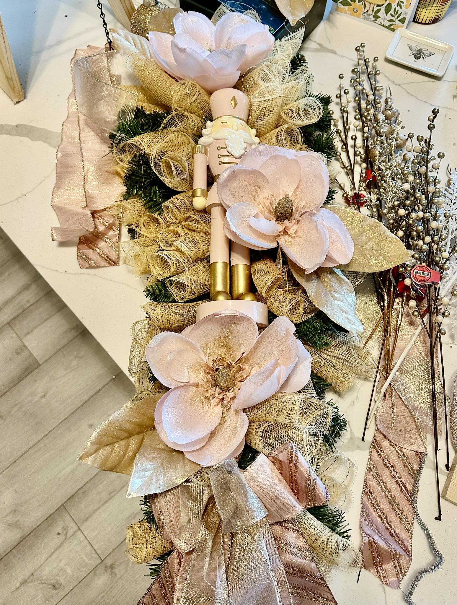 Decorative holiday arrangement with artificial pink magnolia flowers, gold mesh ribbons, a nutcracker figure, and various metallic and ribbon ornaments on a white surface.