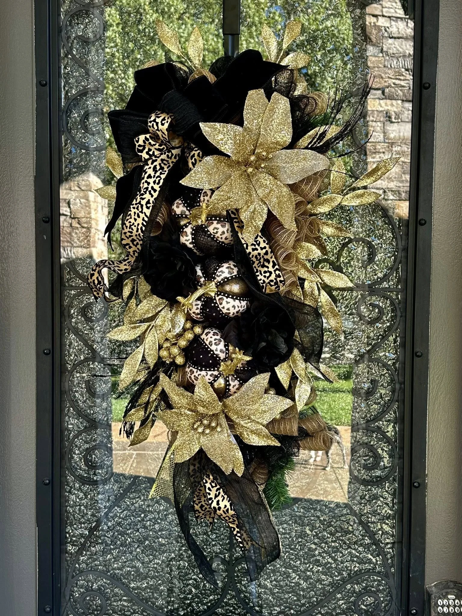 Decorative wreath with large gold poinsettias, black and leopard print ribbons, gold berries, and black mesh, hanging on a glass door with a view of trees and a brick wall outside.