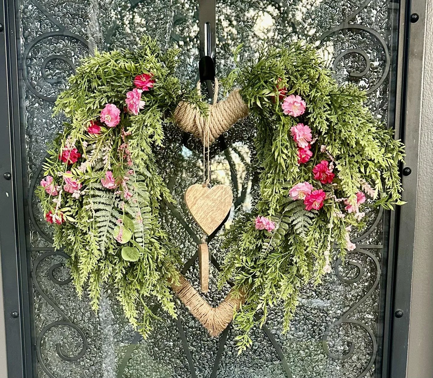 A heart-shaped floral wreath with pink flowers and green leaves, decorated with a wooden heart and a wooden arrow on a glass door with a metal scrollwork design.