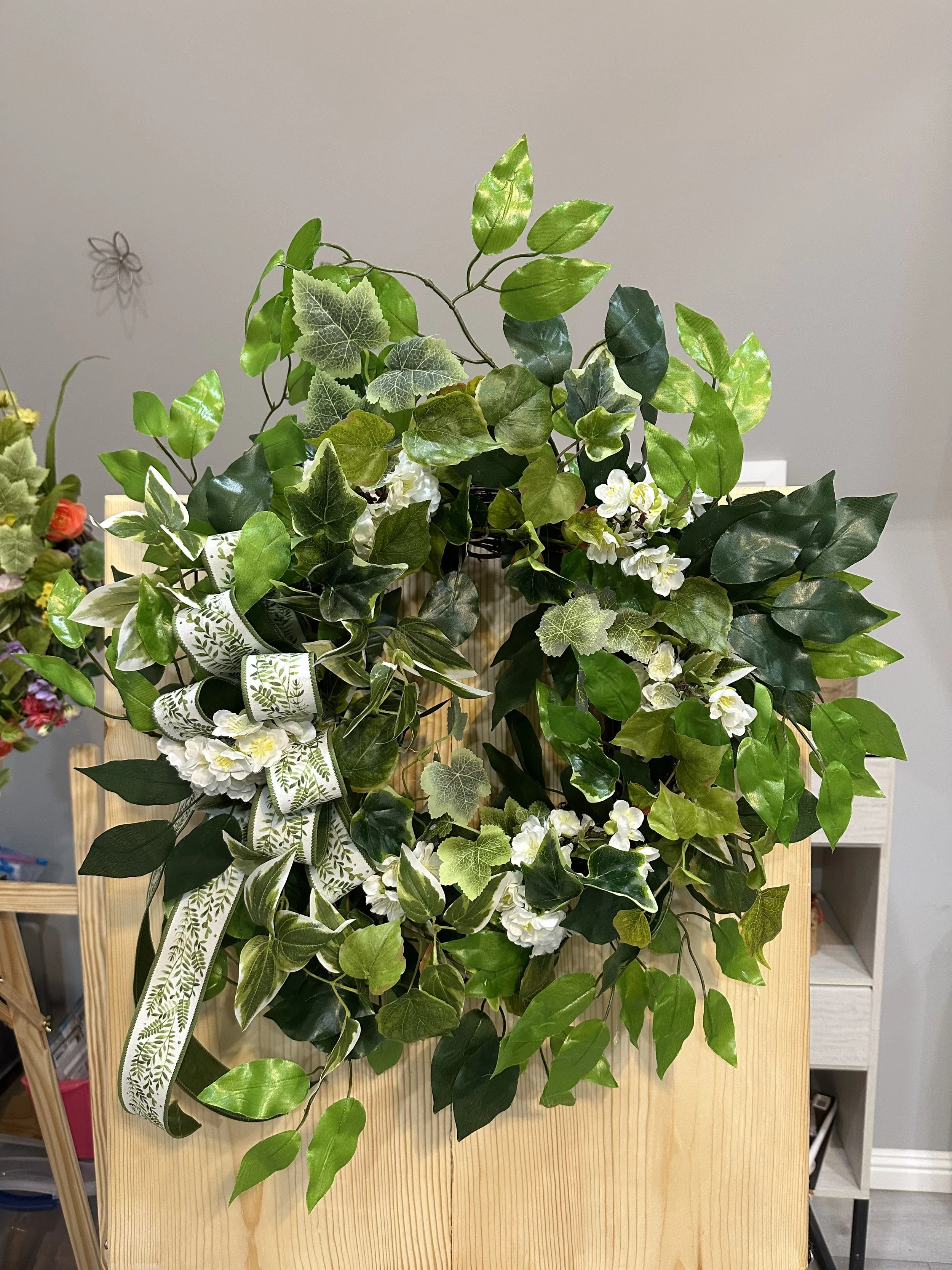 A decorative arrangement of various green plants and ivy with white flowers, adorned with a ribbon, placed on a light-colored wooden stand.