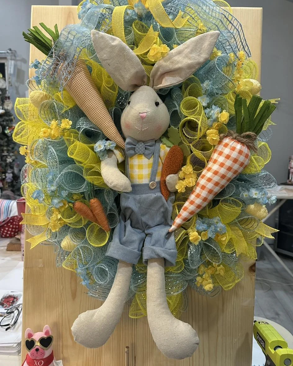 Decorative bunny doll with long ears, wearing a checkered shirt, bow tie, and suspenders, surrounded by a colorful wreath of ribbons, flowers, and large fabric carrots.