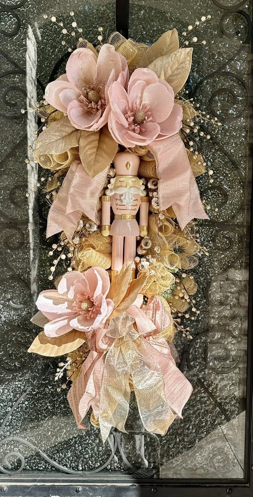 Decorative door wreath with pink flowers, gold leaves, pink ribbons, pearl accents, and a nutcracker figurine at the center, on a glass door with rain droplets.