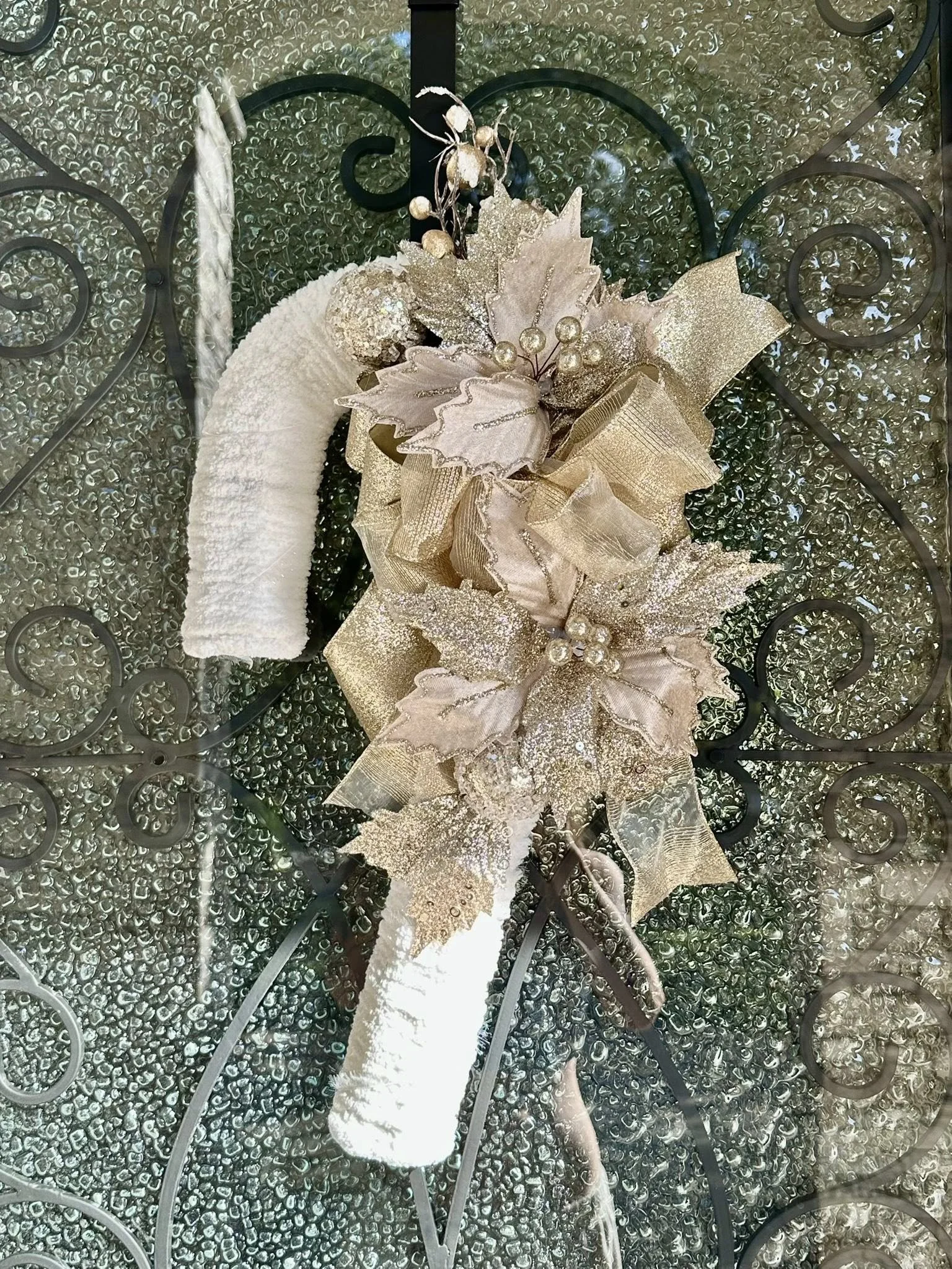 A decorative Christmas wreath with gold and beige poinsettia flowers, glittery leaves, gold accents, ribbon, and berries attached to a textured white spray. It is hanging on a wrought iron decorative gate.