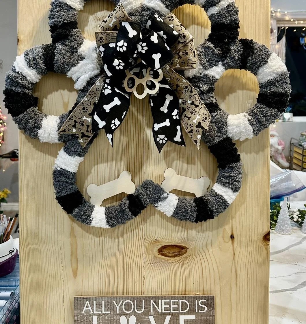 A decorative wreath made of black, gray, and white fuzzy material with a large bow featuring dog bone and paw print patterns. The wreath is mounted on a wooden board with a small sign at the bottom that says, "All you need is".