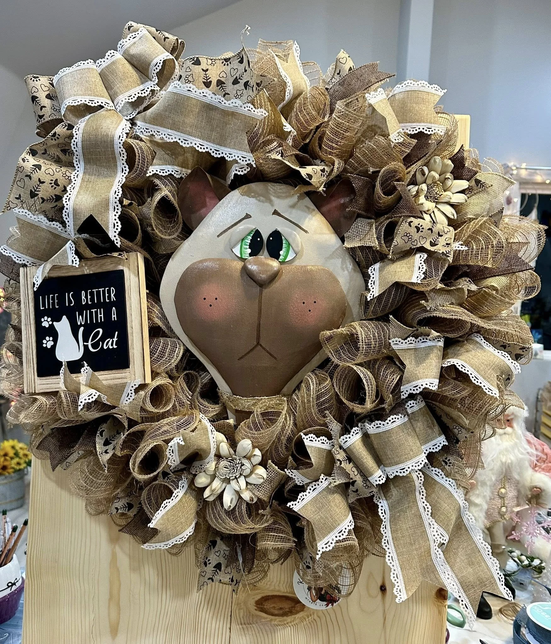 Decorative wall hanging featuring a cat face made of mixed materials surrounded by a large, ruffled, beige and brown ribbon wreath with lace accents, and a small sign that reads "Life is better with a cat" with a cat silhouette.
