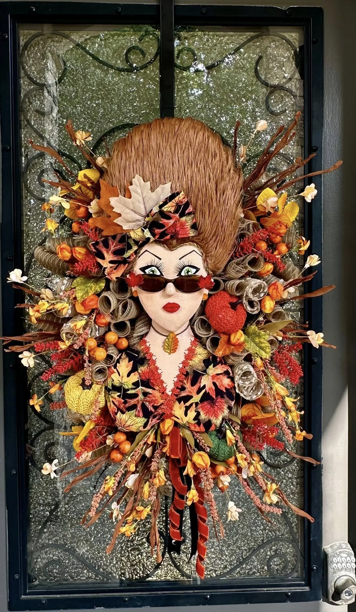 Fall-themed door decoration featuring a whimsical woman with high hair made of orange and gray curls, wearing sunglasses, red lipstick, and surrounded by autumn leaves, berries, apples, and flowers.