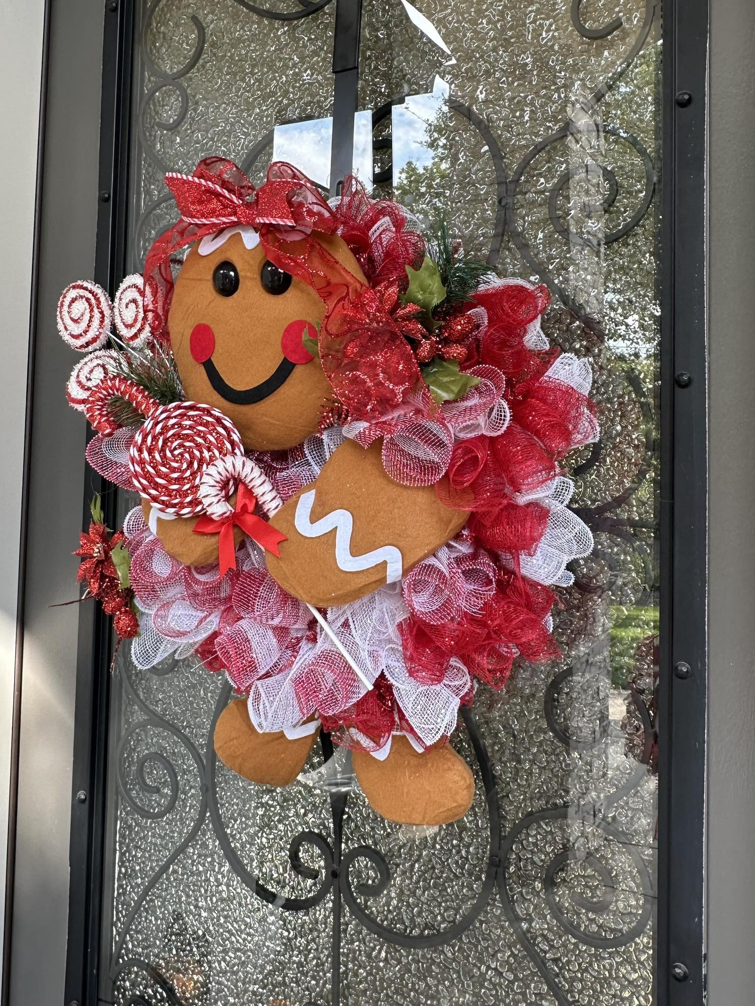 Decorative gingerbread man holiday wreath hanging on a door with glass and wrought iron design. The gingerbread man has a smiling face, black eyes, and red cheeks, and is surrounded by red, white, and green holiday ribbons, bows, and holly leaves.