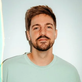 Portrait of a young man with short brown hair, a beard, and wearing a light-colored shirt, standing against a plain light background.