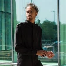 A man with curly hair and a beard standing inside a building near large glass windows, wearing a black hoodie and holding a small object in his hands
