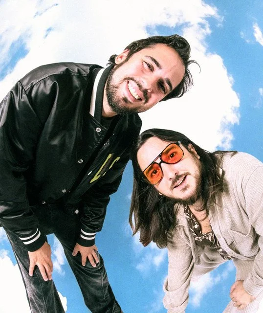 Two men posing against a blue sky with clouds, smiling and leaning toward the camera
