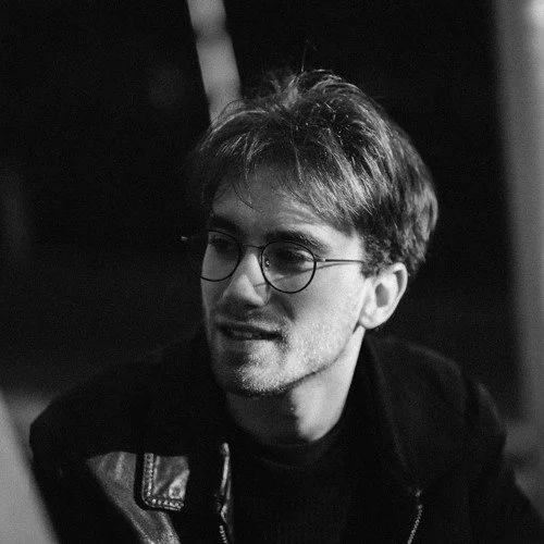 A young man with glasses and light-colored hair, smiling and looking to the side in a black and white photo.