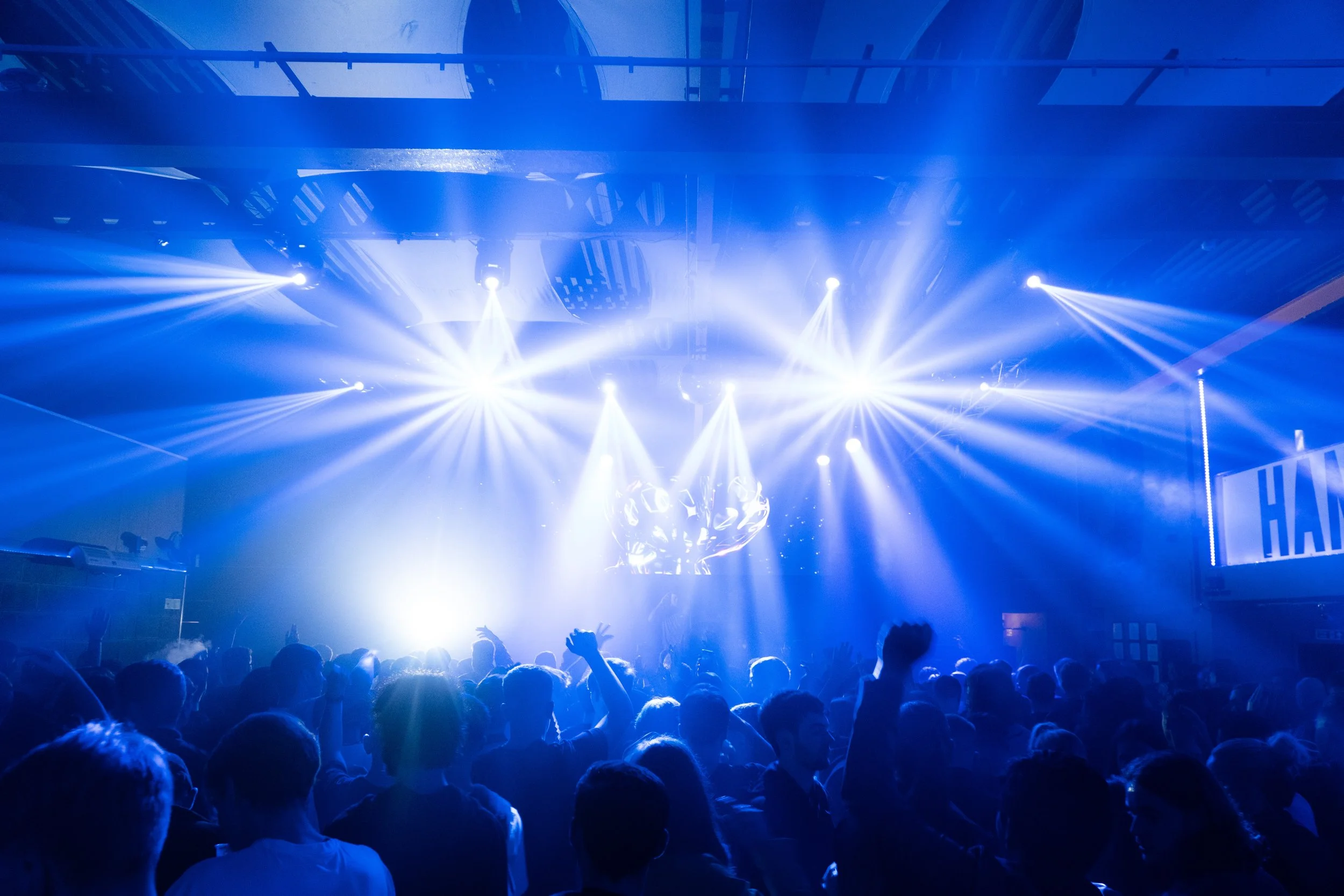 Crowd of people at a concert with blue and white stage lights and a DJ or performer on stage.