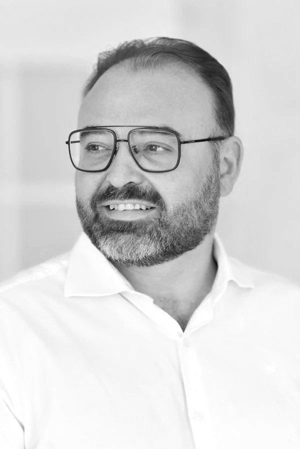 Hombre con barba, gafas y camisa blanca, sonriendo en un entorno interior en blanco y negro.