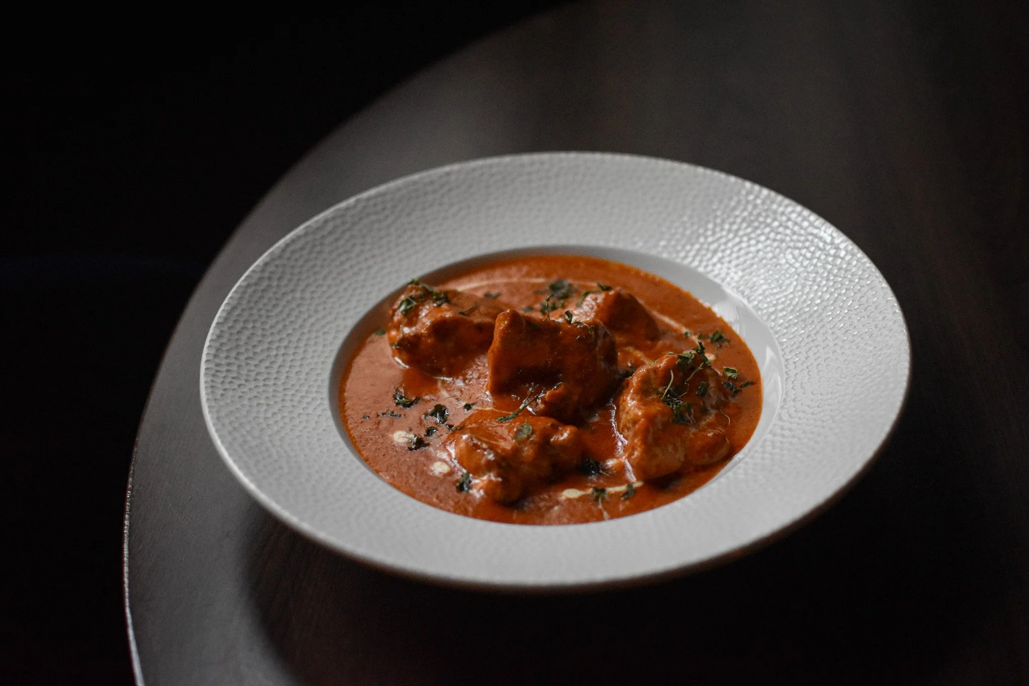 A white textured bowl filled with Butter chicken which is a tomato-based sauce, garnished with chopped herbs, on a dark surface at Nomi restaurants
