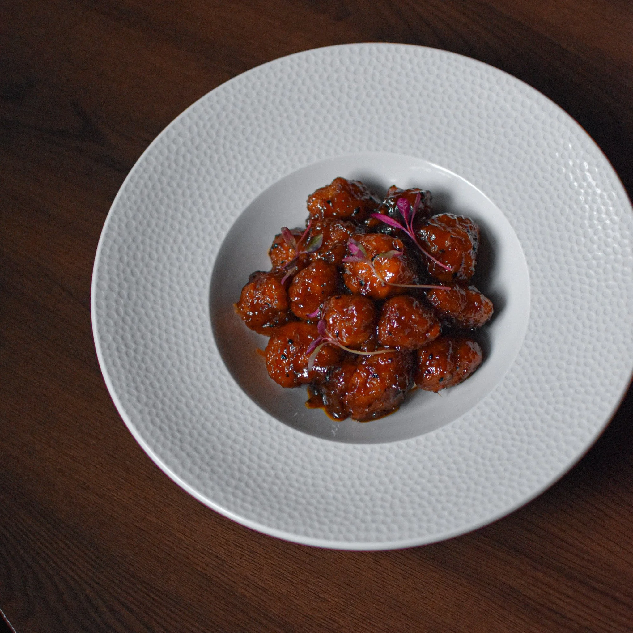 Plate of mango maple chicken  coated in glossy, dark sauce, garnished with small purple microgreens on a white textured plate, on a wooden surface at nomi restaurants, its the best seller