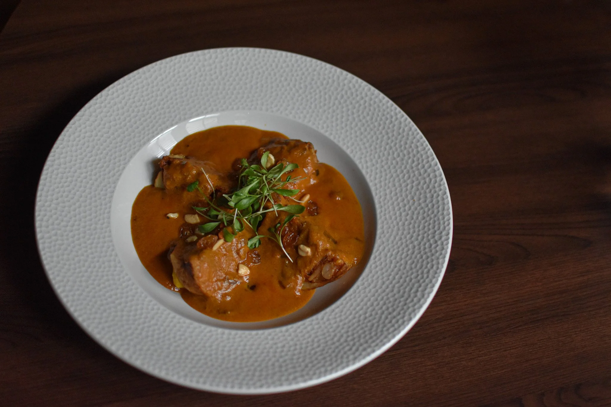 A white textured bowl with stuffed potatoes curry garnished with microgreens and chopped nuts on a dark wooden table at Nomi Restaurants