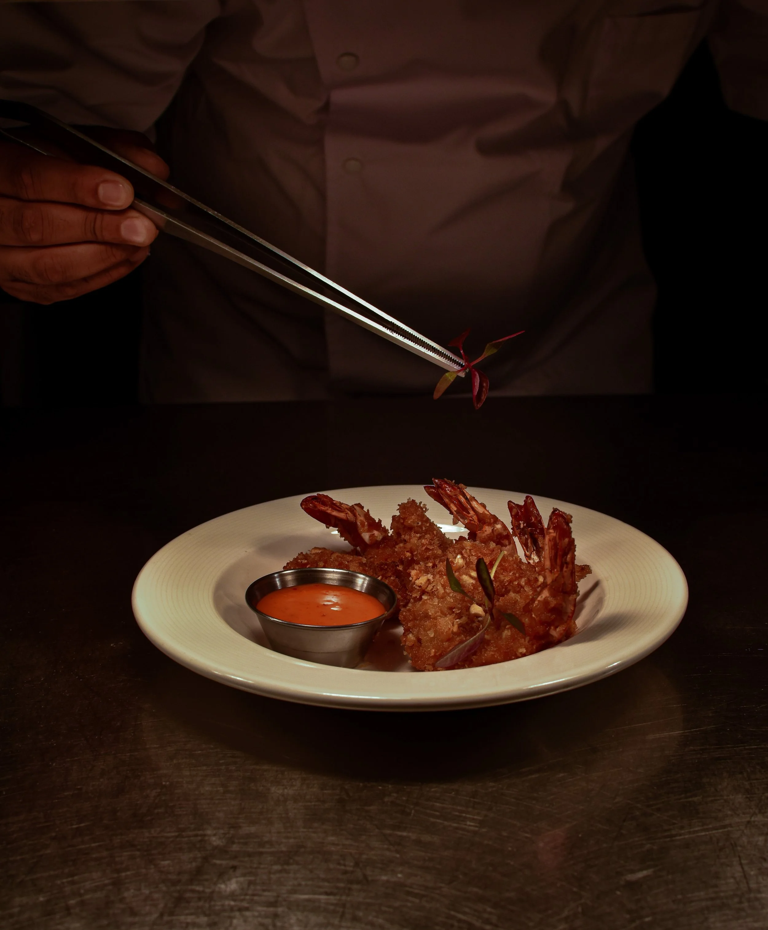 Head chef of Nomi Restaurants in a white shirt garnishing a plate of fried shrimp with herbs, with a side of dipping sauce on a white plate.