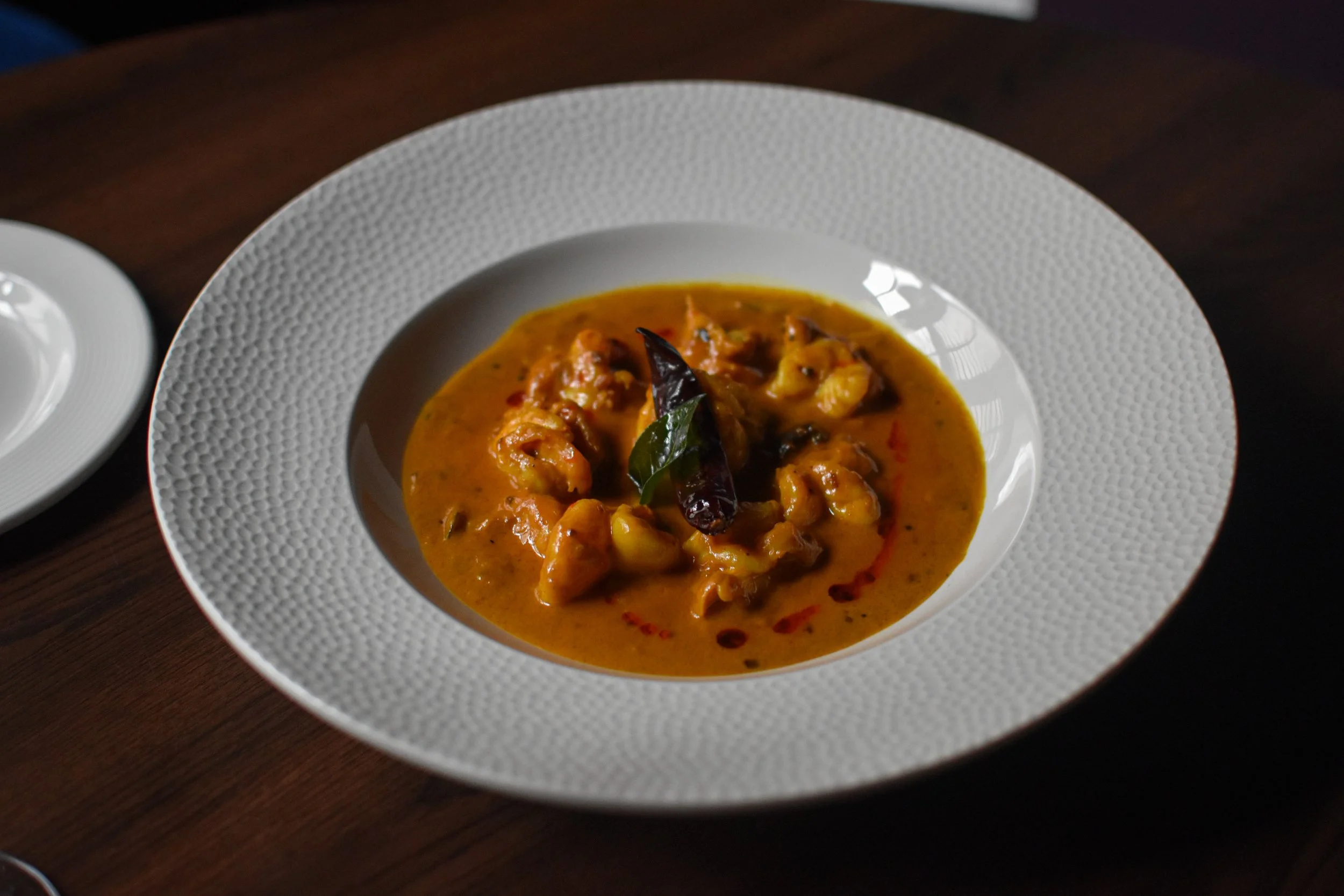 A white bowl with a textured rim containing a serving of yellow curry with juicy prawns and spices, garnished with a dried chili and a curry leaf, on a dark wooden table at Nomi Restaurants