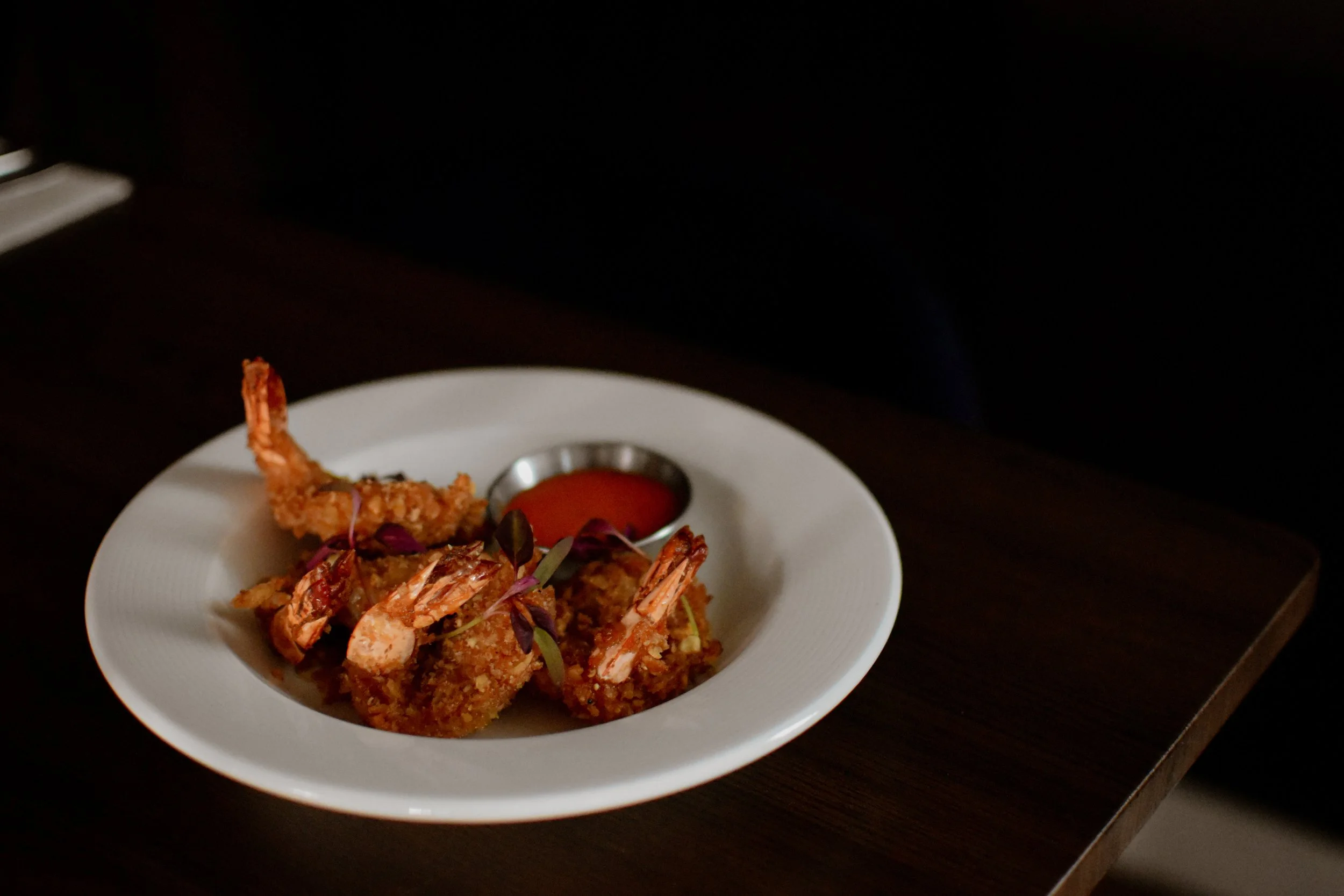 Plate of cashew crusted prawns with dipping sauce on a dark wooden table at Nomi Restaurants