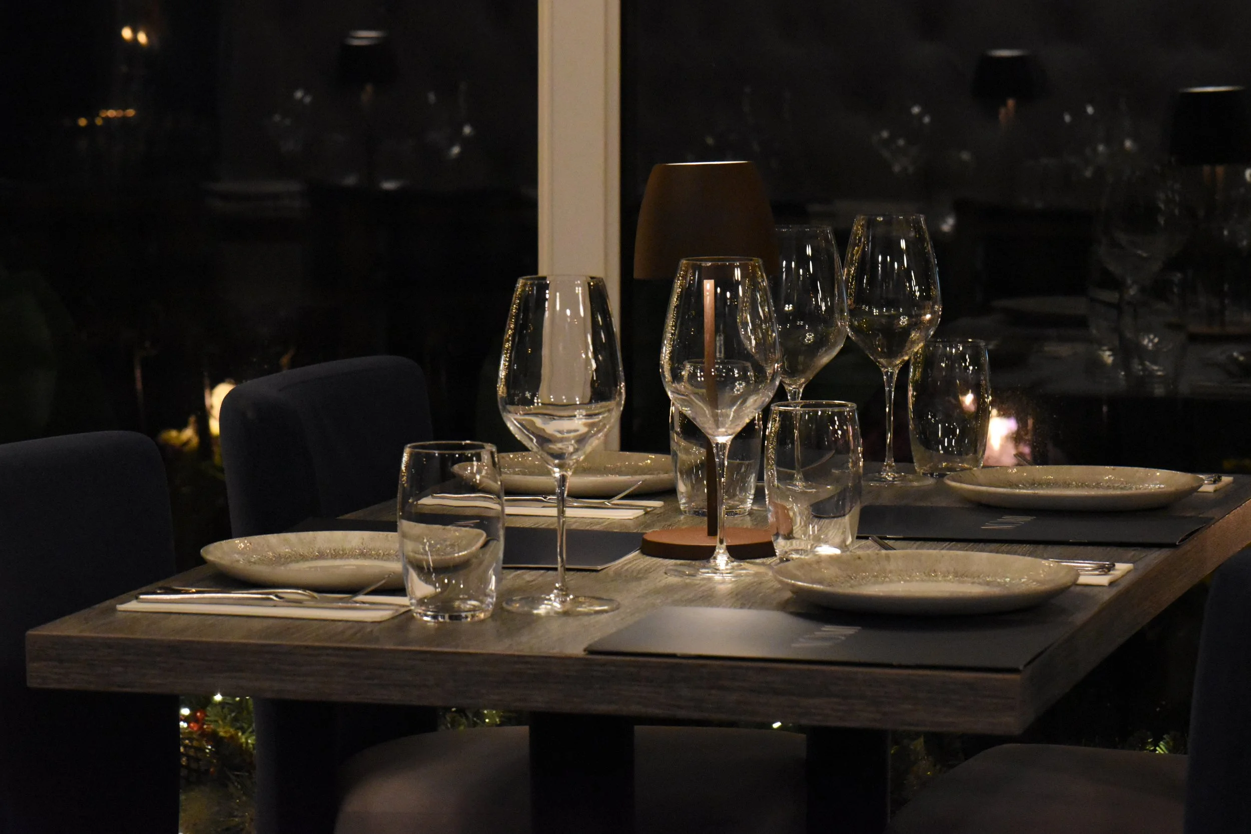 A dining table set with empty plates, wine glasses, water glasses, and silverware, with a dark background and warm lighting at Nomi Bangor overlooking the bangor pier