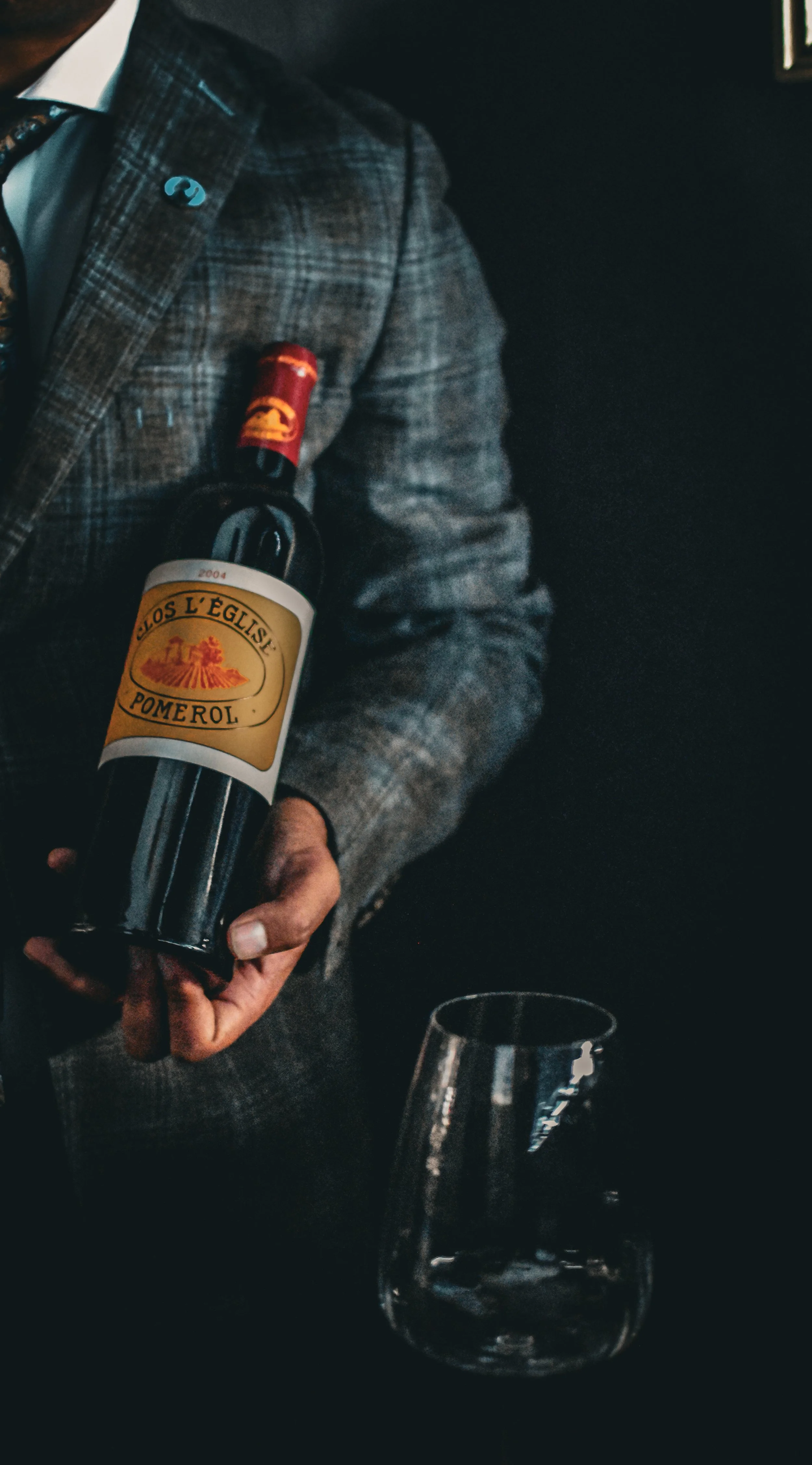 Person in a plaid blazer holding a bottle of Los L' Eglise Pomerol wine in a dark room, with an empty glass on a table in front at Nomi Restaurant