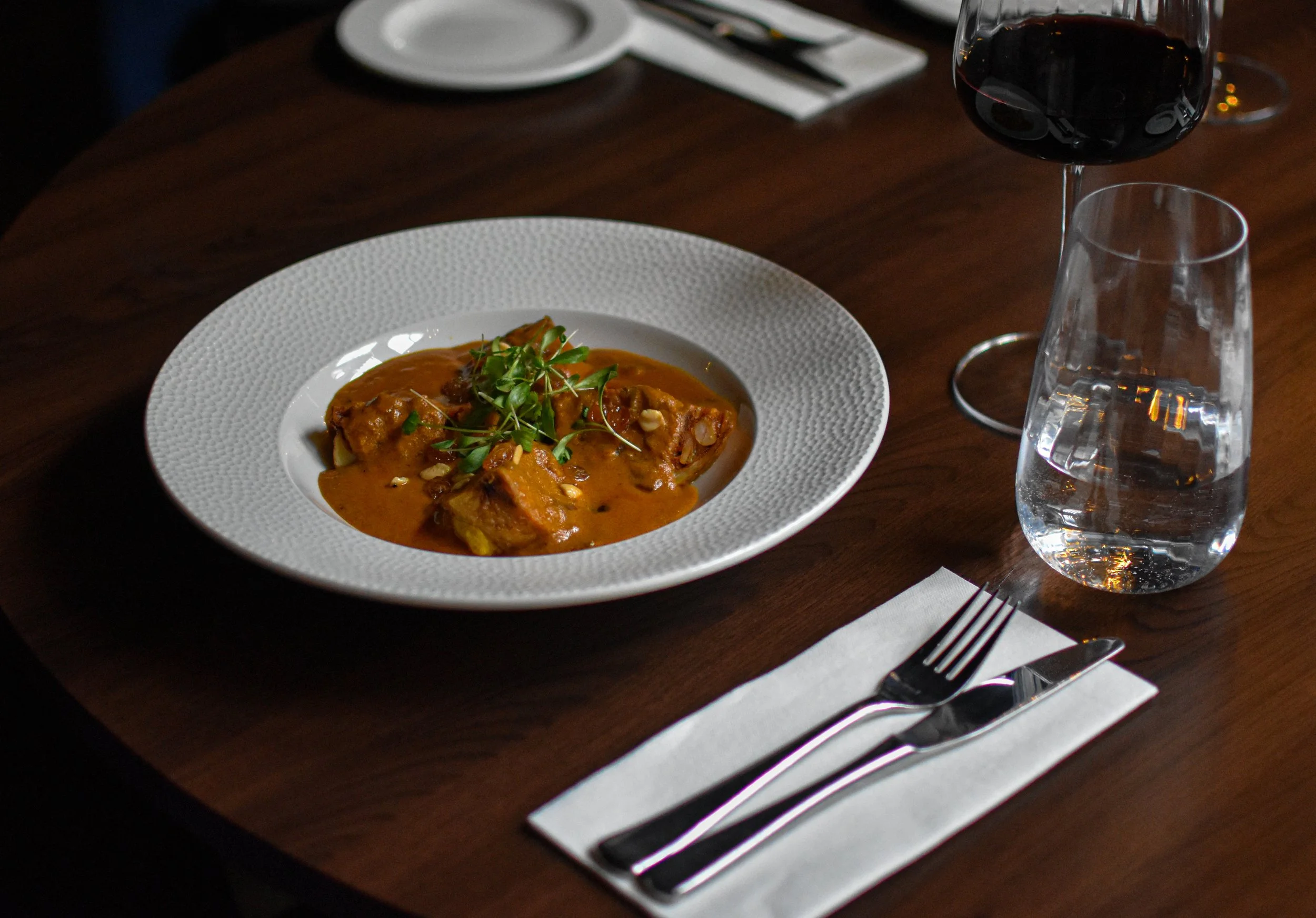 A white textured plate with a serving of stuffed potatoes garnished with microgreens, placed on a dark wooden table with a glass of red wine, a water pitcher, and utensils wrapped in a white napkin at Nomi Restaurants