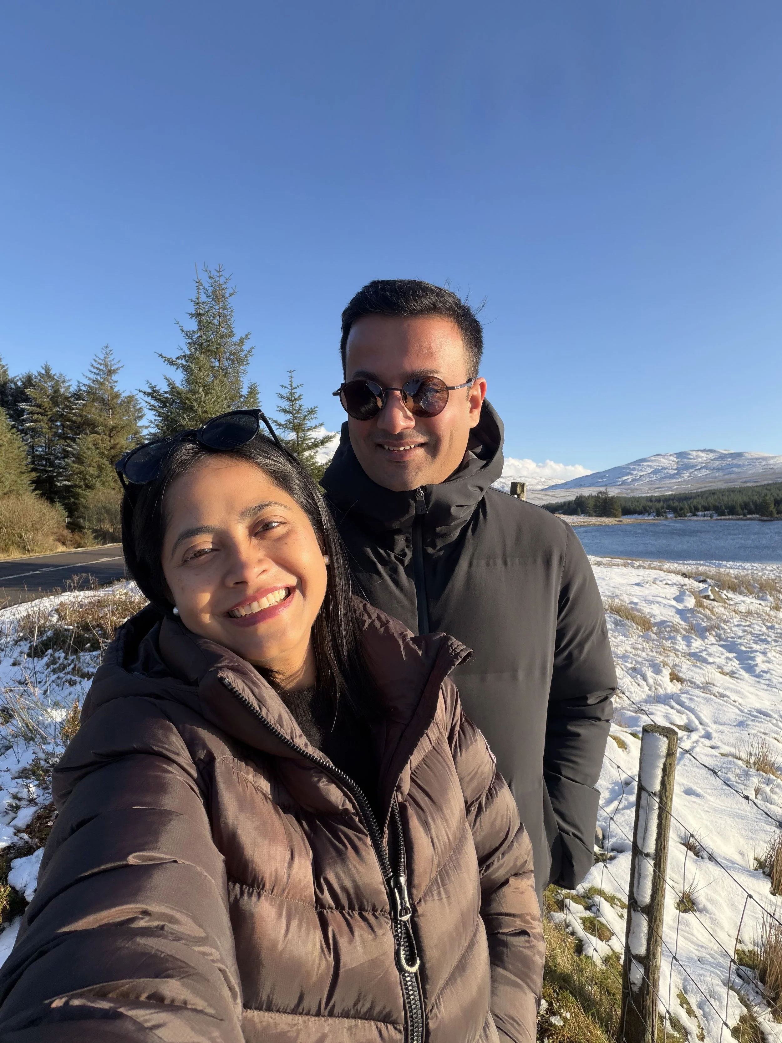 A smiling woman is Nomi and man dressed in winter jackets and sunglasses is her husband and the head chef of Nomi restaurant  taking a selfie outdoors near a lake with snow-covered ground, trees, and mountains in the background on a sunny winter day.