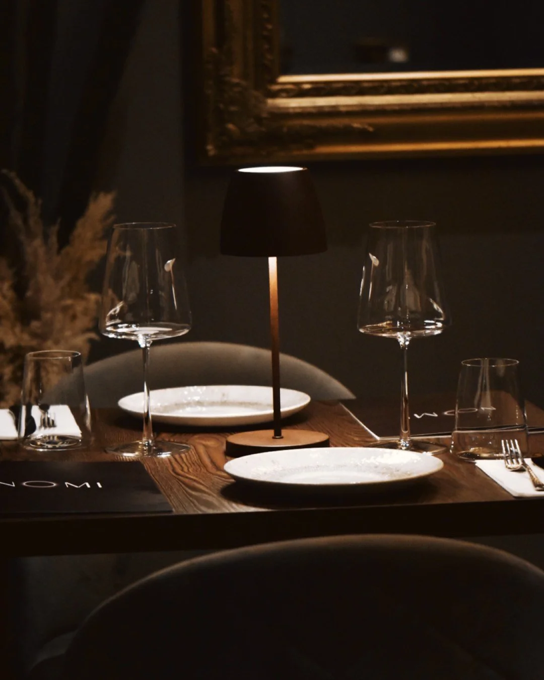 A table setting in a dimly lit Nomi restaurant at Nomi Porthmadog with a modern black table lamp, white plates, four wine glasses, and a complete silverware set.