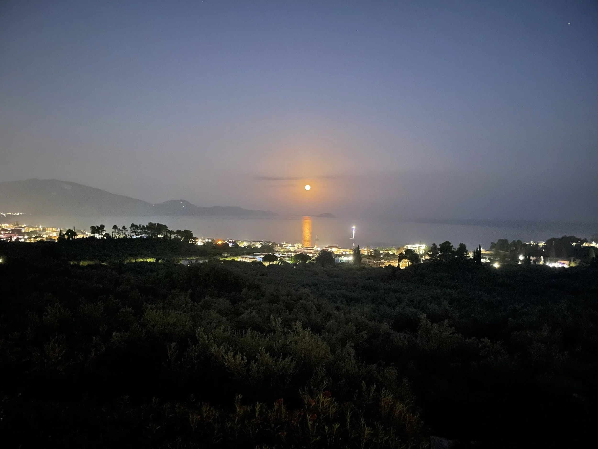 Nighttime landscape with a full moon over a coastal city, with the moon's reflection on the water, and mountains in the background.