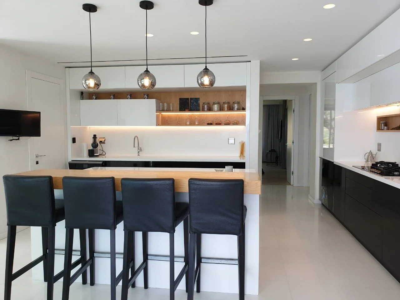 Modern kitchen with black and white cabinetry, black bar stools, wood countertop, open shelving with jars, and pendant lights.