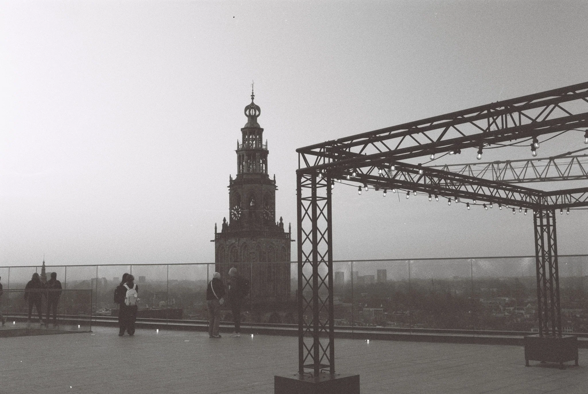 People standing on a rooftop with a city skyline and a tall historic clock tower visible in the background, under an overcast sky.