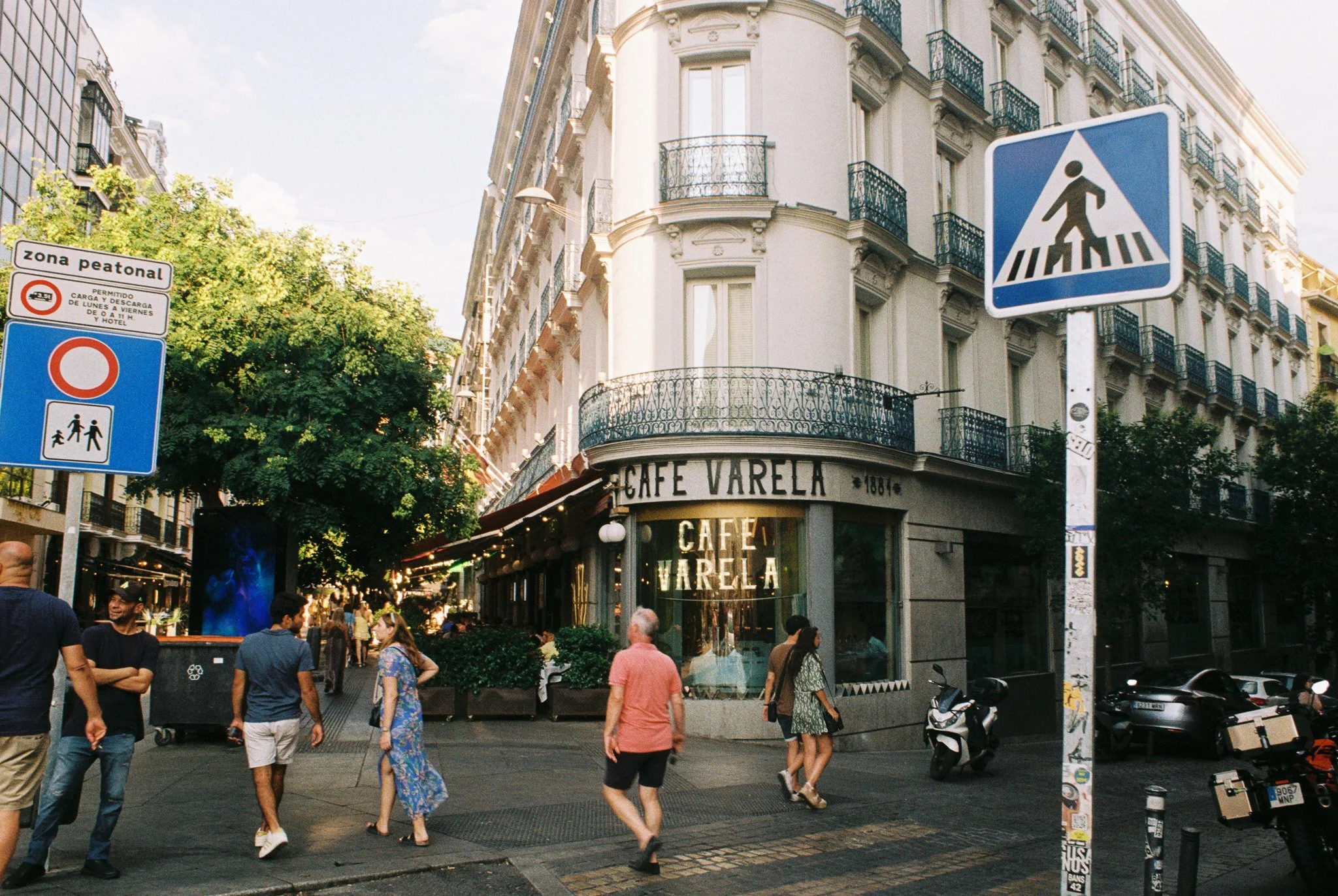 A busy city street corner with pedestrians walking. A corner building has a sign that reads 'Café Varela'. There are various street signs, including a pedestrian crossing sign and a zone sign indicating restricted access.