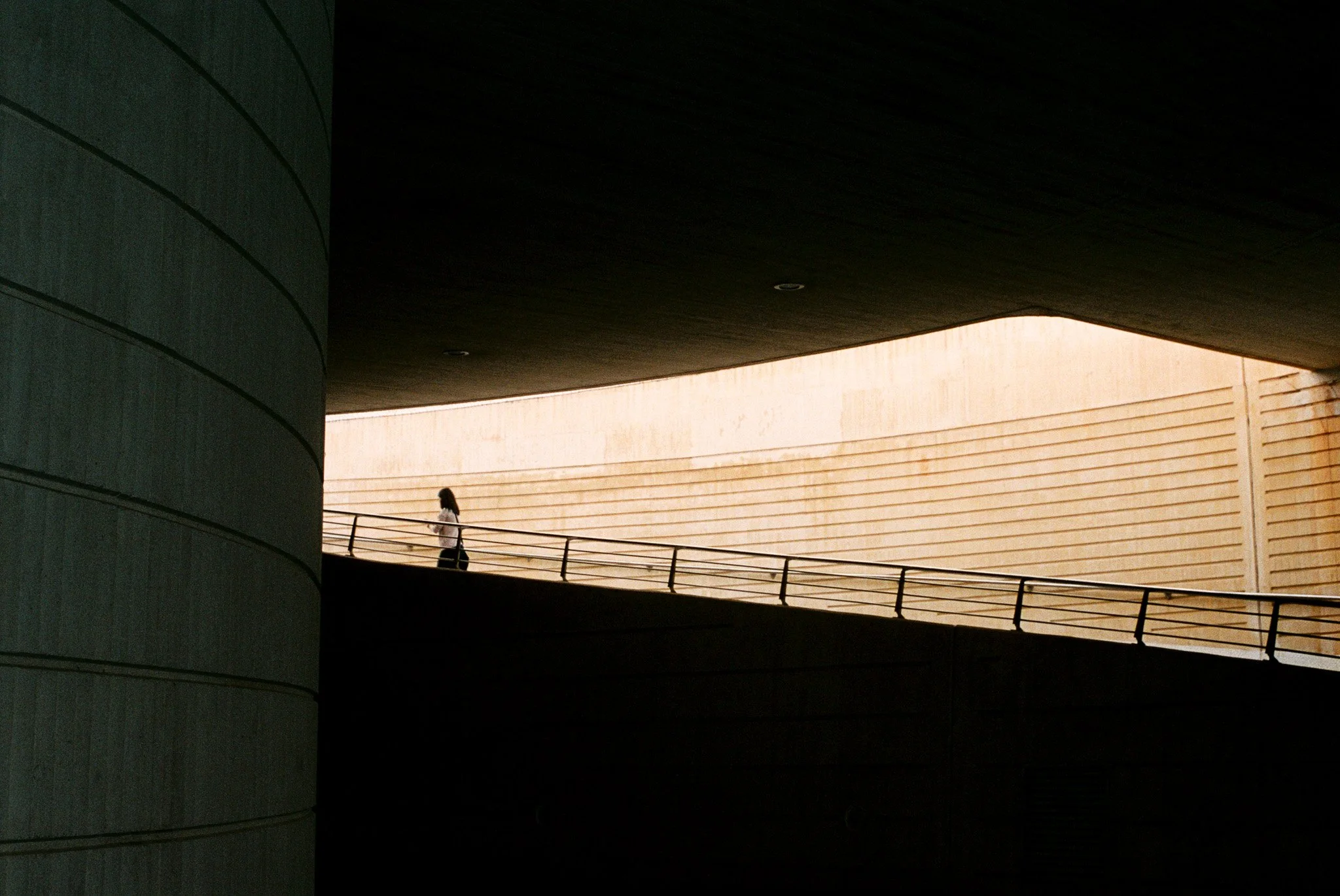 A woman walking along a curved, elevated walkway with a metal railing, underneath a large overhanging dark ceiling, with a beige wall in the background.