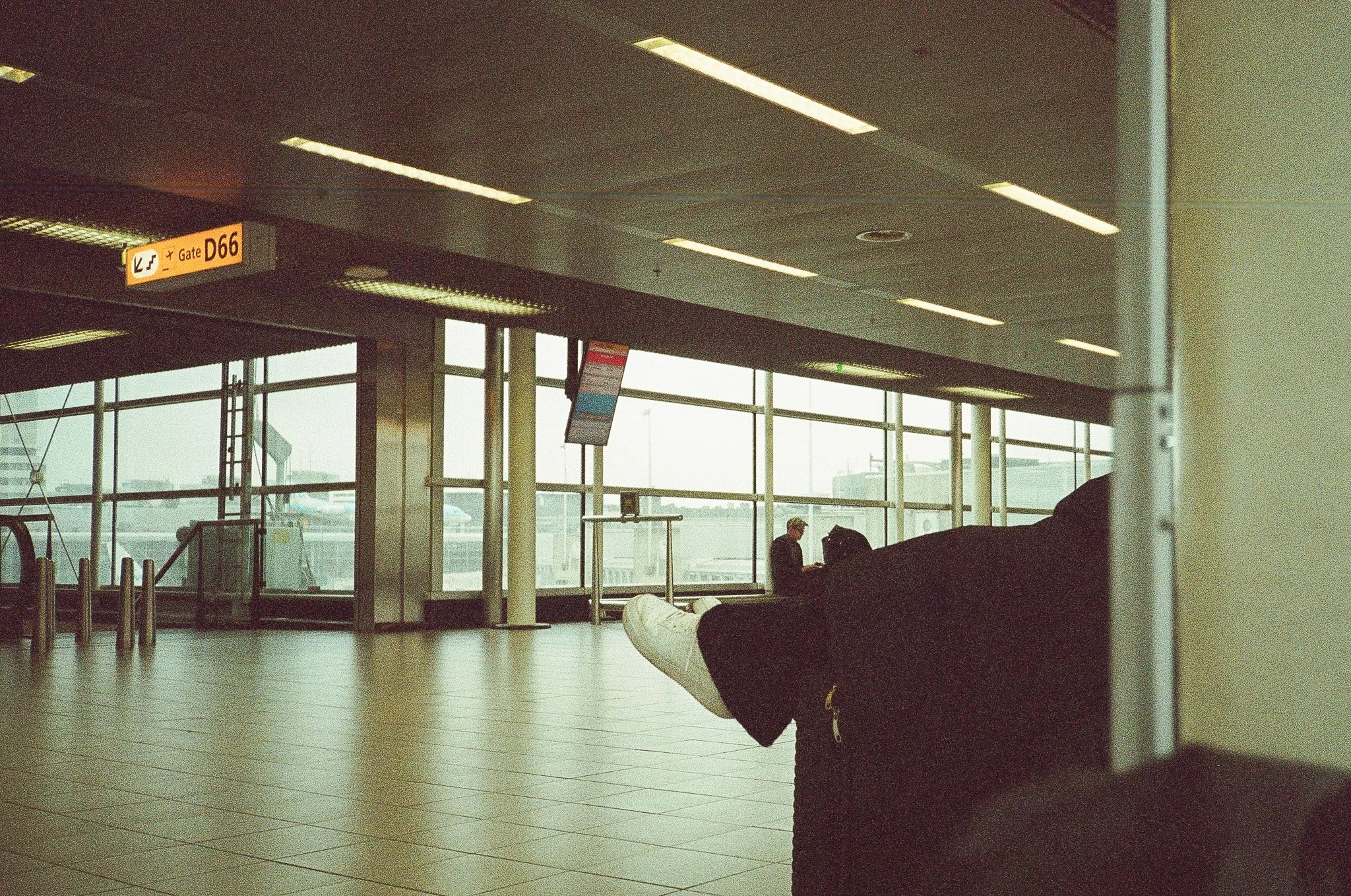 Empty airport terminal with a few passengers, including one sitting with sitting with their legs crossed, and large windows showing airplanes outside.