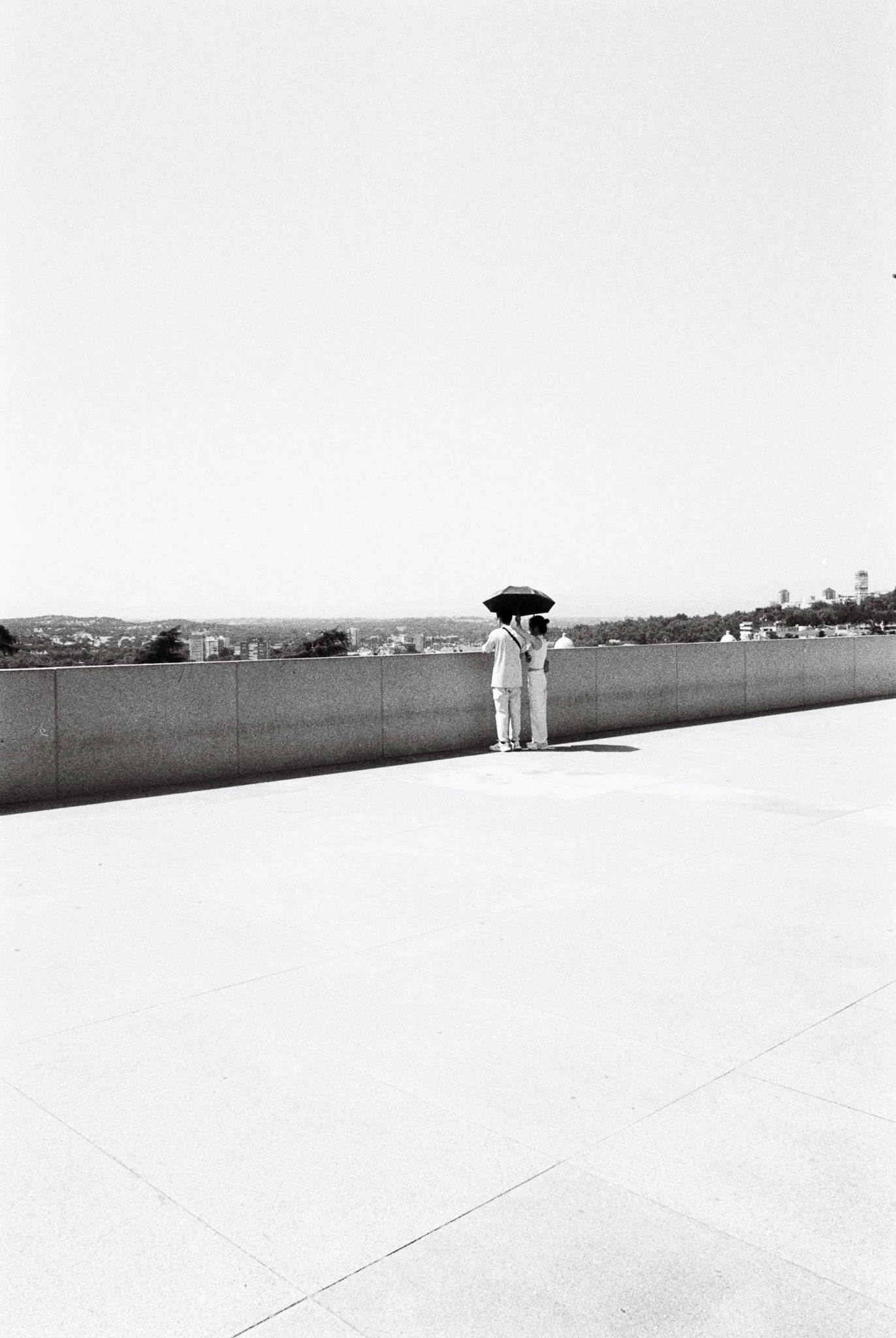 Two people standing close under an umbrella on a rooftop, overlooking a cityscape.
