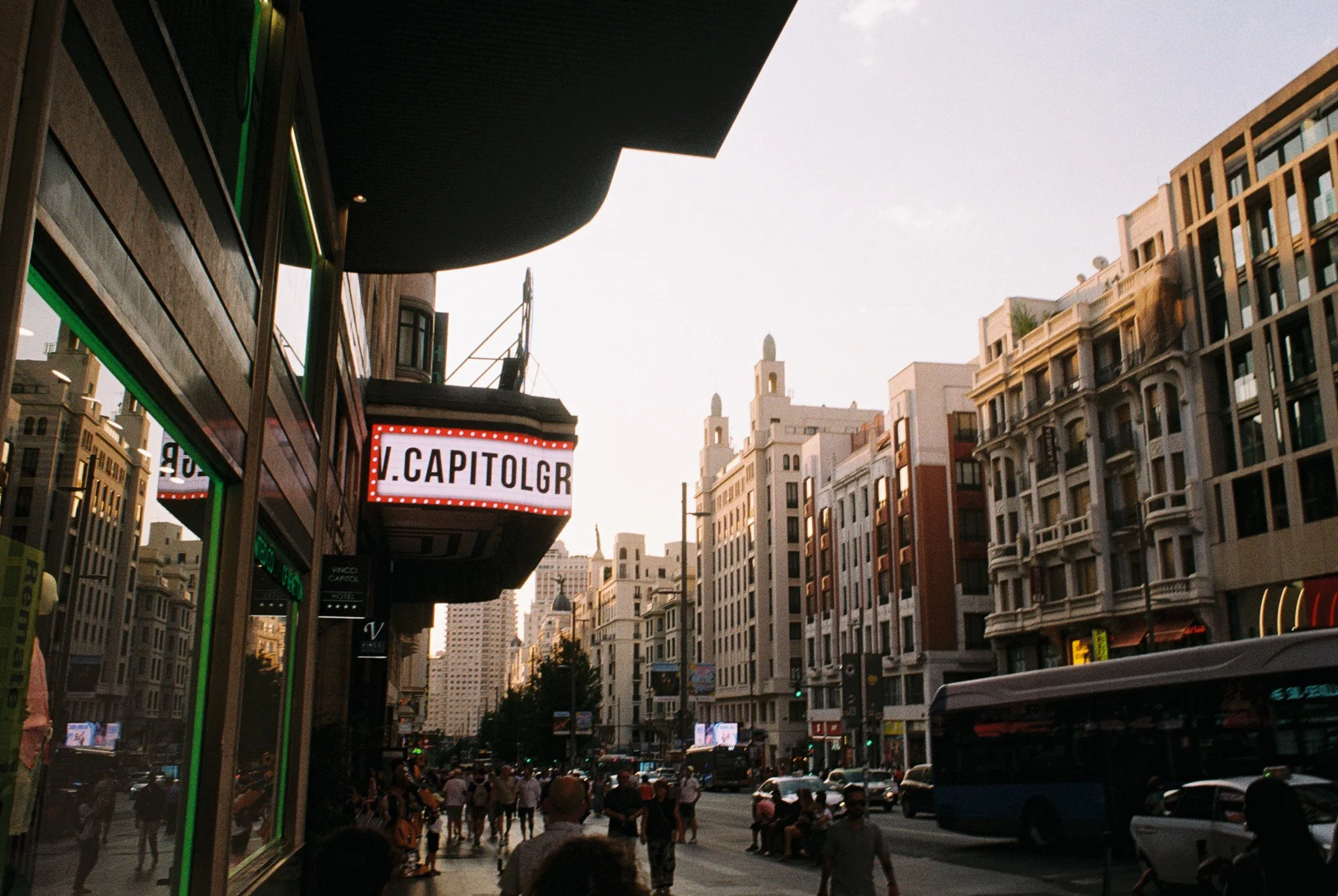 City street scene featuring large buildings with ornate facades, a bus on the road, people walking on the sidewalk, and an electronic marquee sign that reads 'V. CAPITOLGR'.