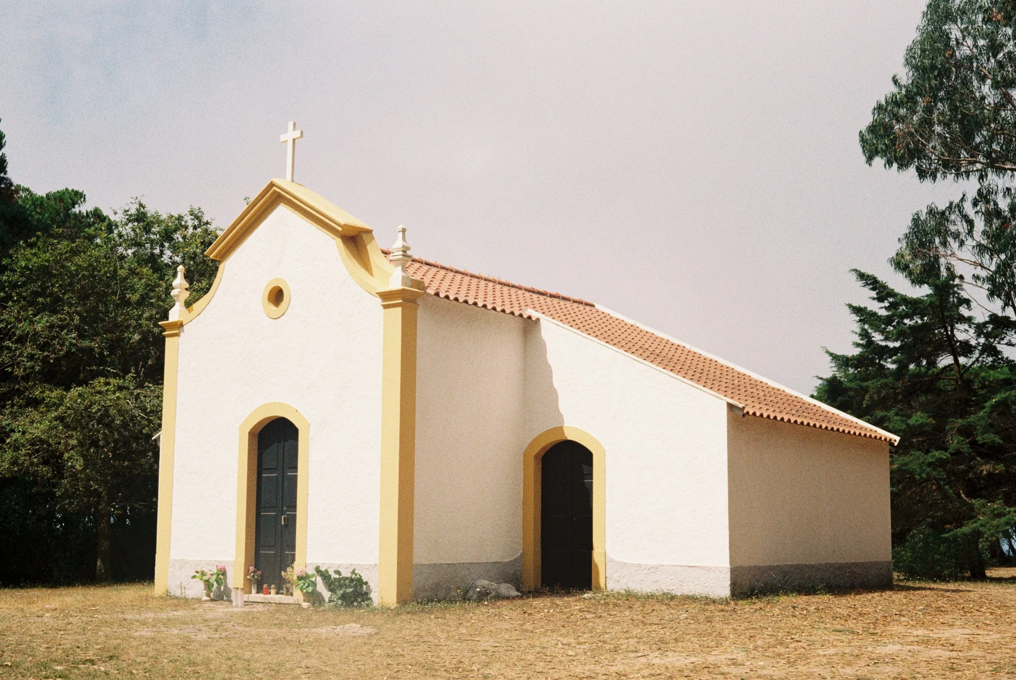 Small white chapel with a cross on top, yellow trim around door and windows, terracotta roof, surrounded by trees and open ground.