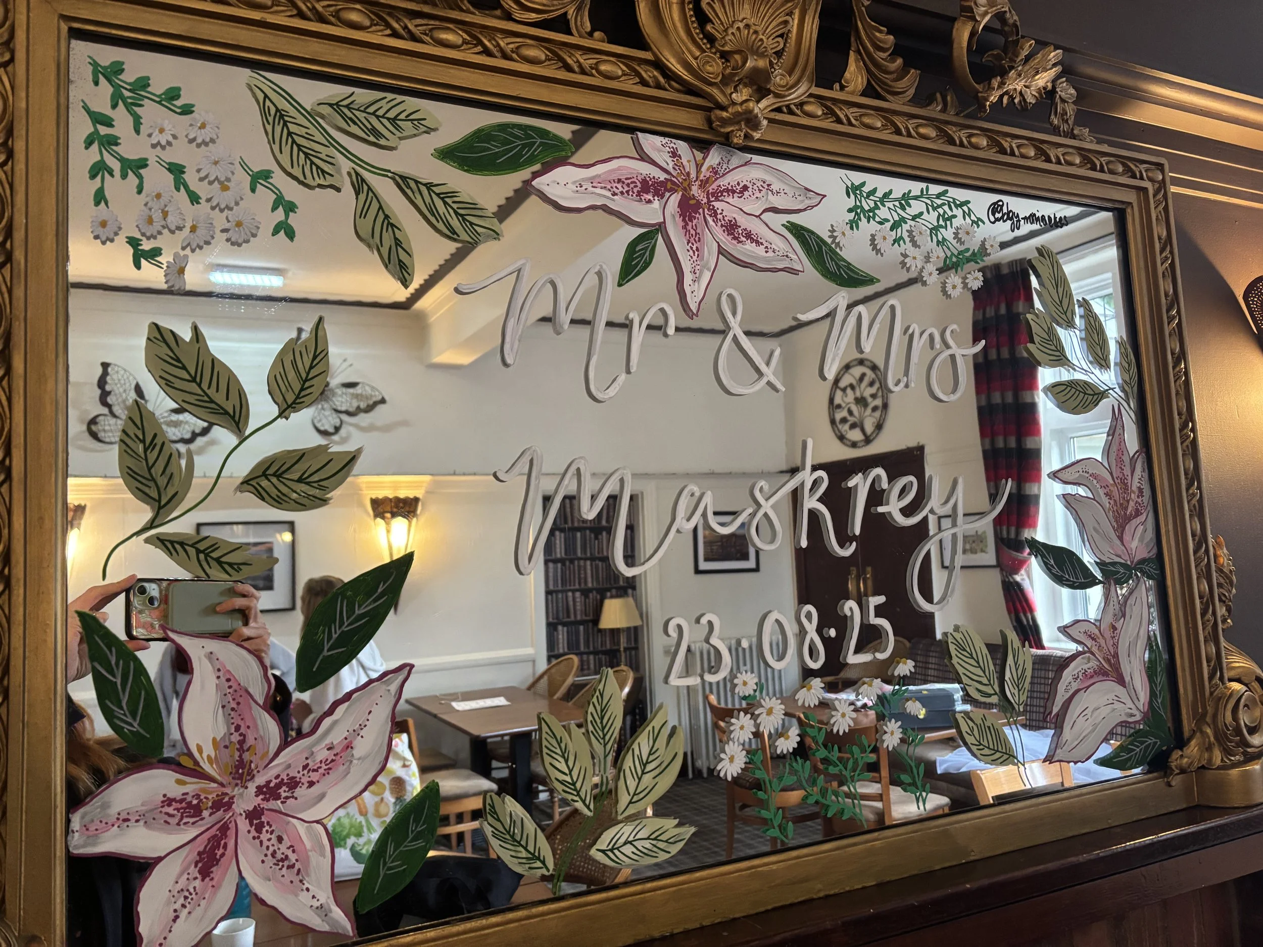 Decorative mirror with floral and leaf designs, inscribed with 'Mr & Mrs Mashrey 23.08.25', reflecting a cozy, well-lit room with furniture, curtains, and framed pictures.