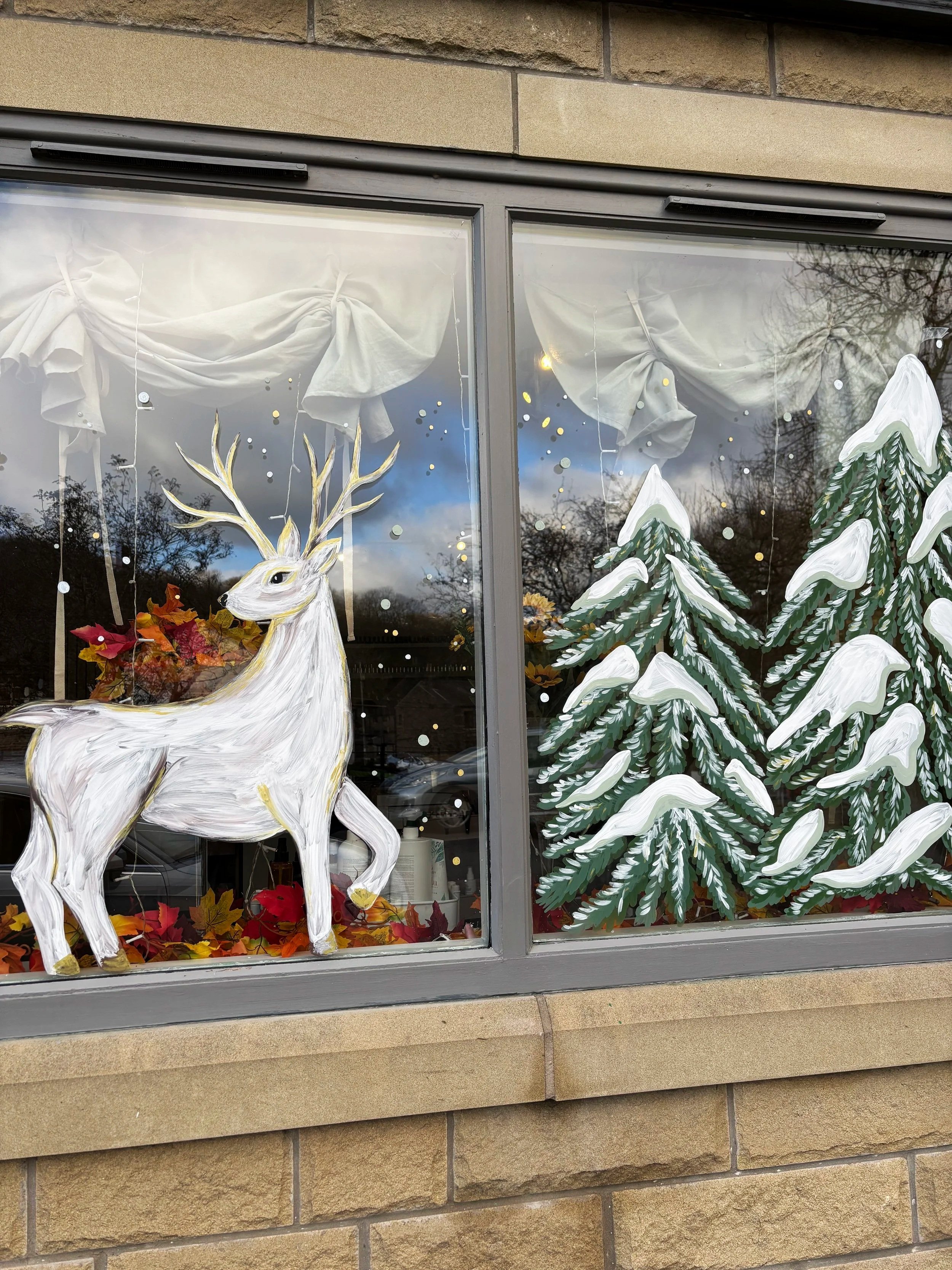 Hand painted christmas window of snow laden pine trees and a reindeer in white and gold. Hand painted christmas window. Painted window display. 