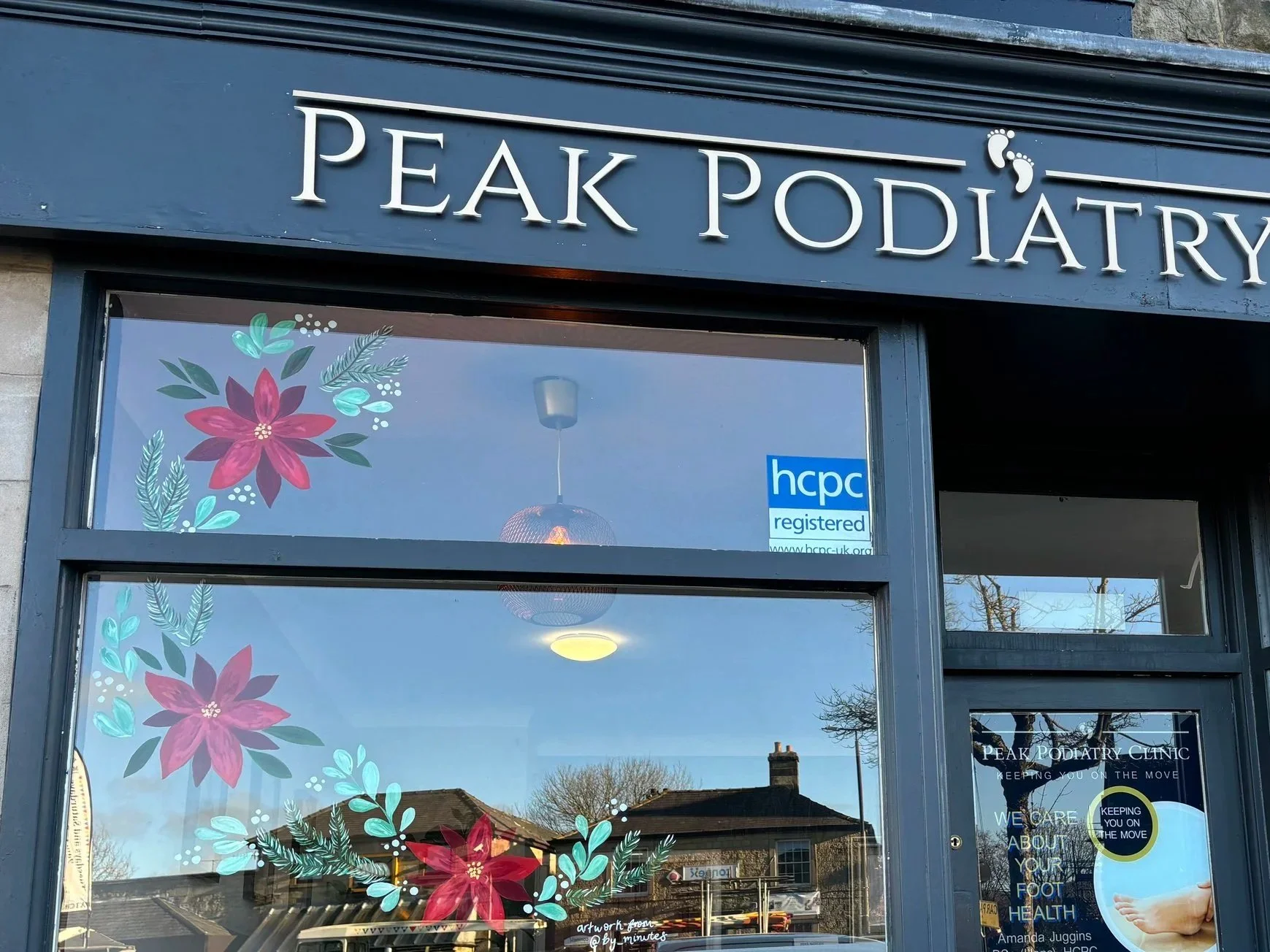 Exterior view of Peak Podiatry Clinic storefront with large windows decorated with pink and green floral decals, blue signage displaying the clinic name, and a door with a poster listing podiatry services.
