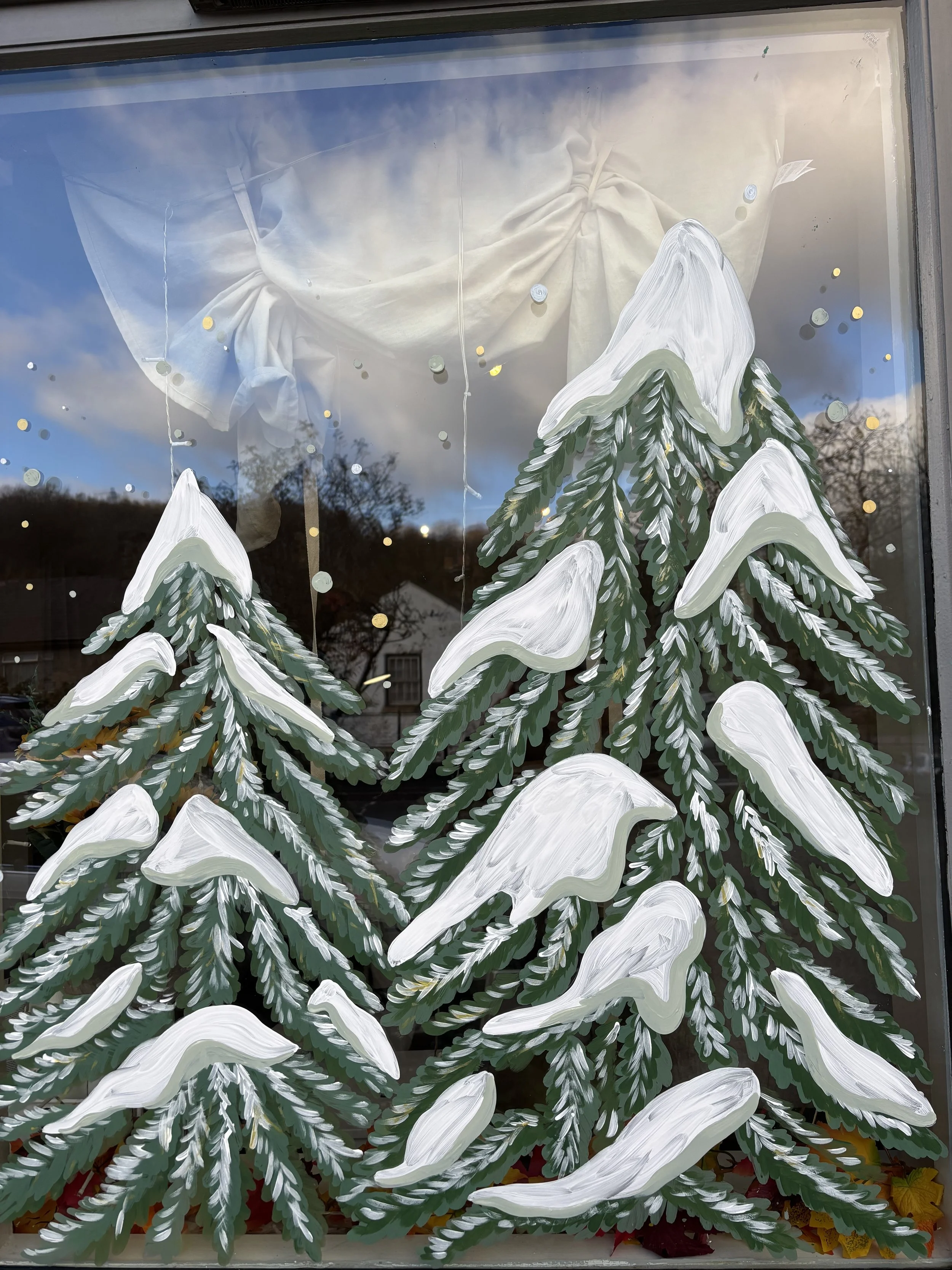 Hand-painted winter scene on window with snow-covered evergreen trees and a background of blue sky and clouds.
