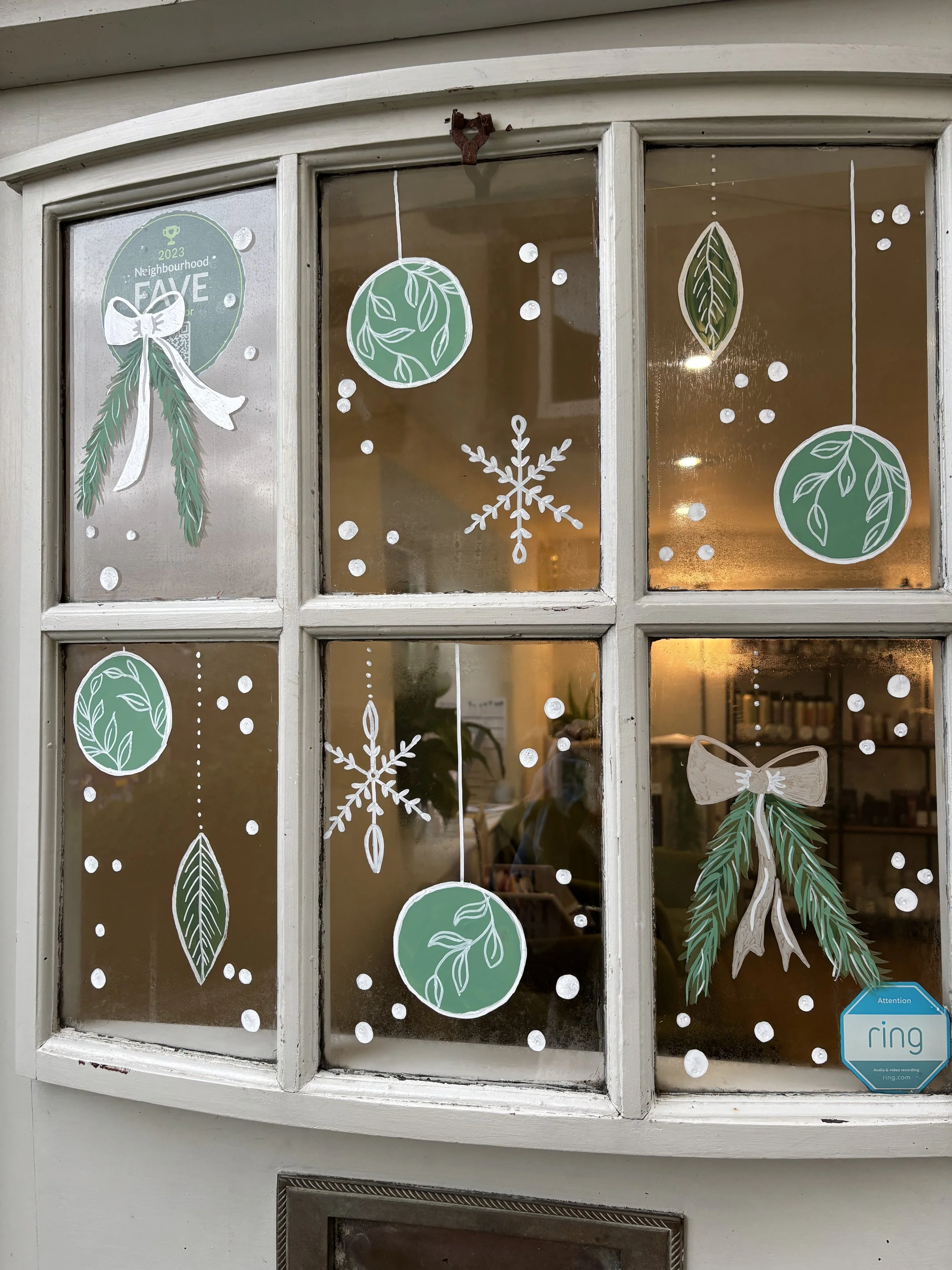 Decorated window with white and green Christmas ornaments and snowflakes, featuring leaves and pine branches, and a 'Neighbourhood Favorite' sticker.