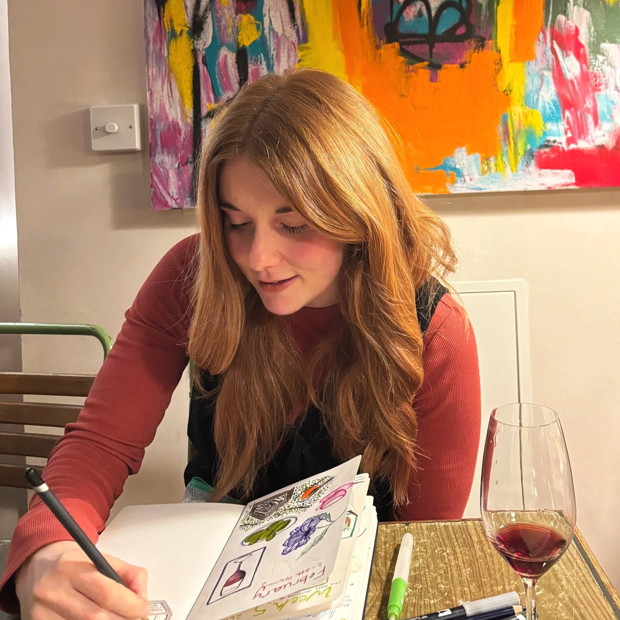 A woman with long, red hair wearing a rust-colored long sleeve shirt and black vest is sitting at a table, drawing or writing in a notebook with various drawings inside. A glass of red wine, a green marker, and a black pen are on the table. Behind her, there is a colorful abstract painting on the wall.
