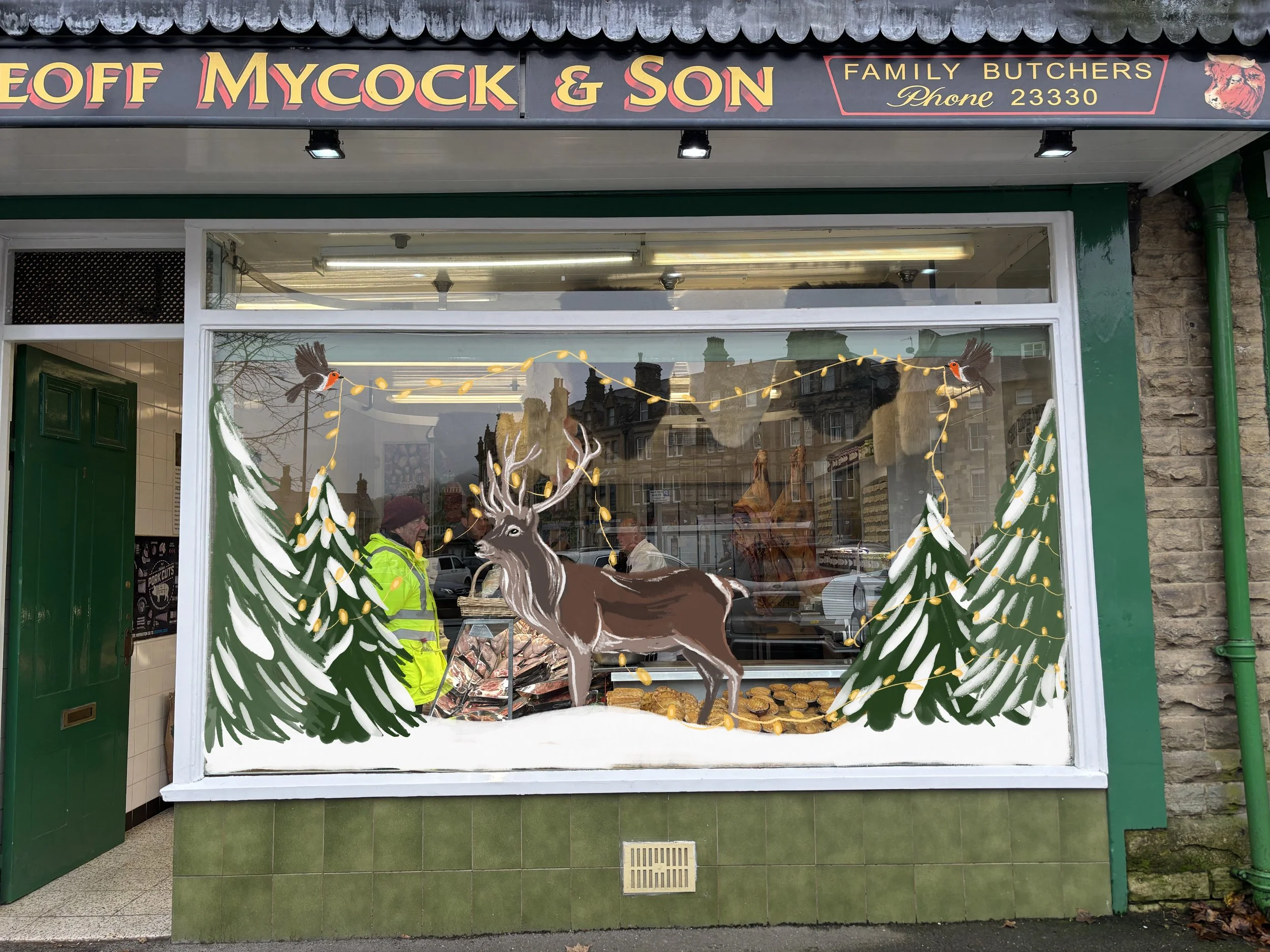 Front window of a butcher shop decorated with Christmas-themed illustrations, including a reindeer, pine trees, and hanging lights, with the shop's sign above.