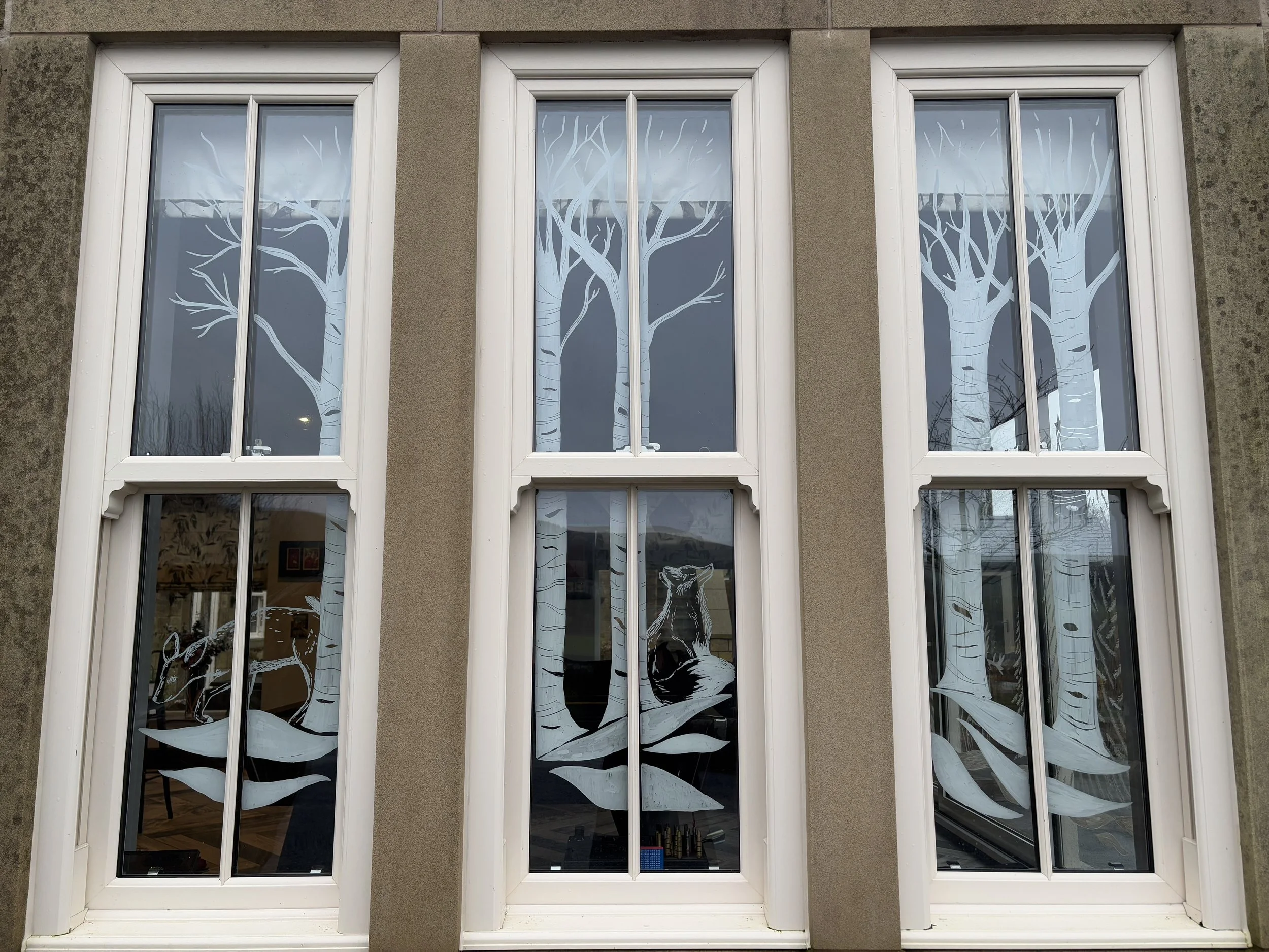 Decorative white window clings depicting trees, leaves, and a sitting fox outside a building with three large rectangular windows.