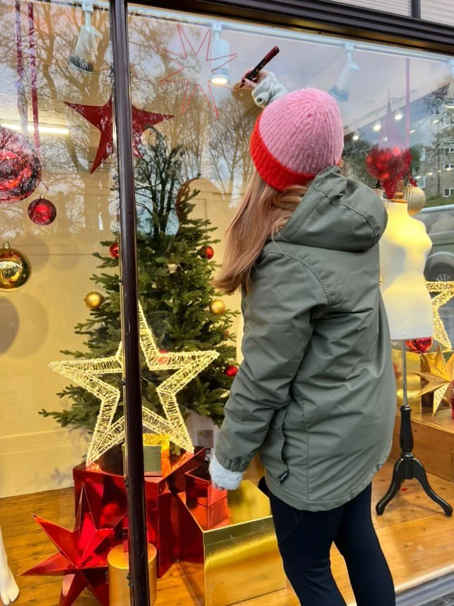 Person in green jacket and pink-red knit hat decorating a Christmas window display with a marker in the store.