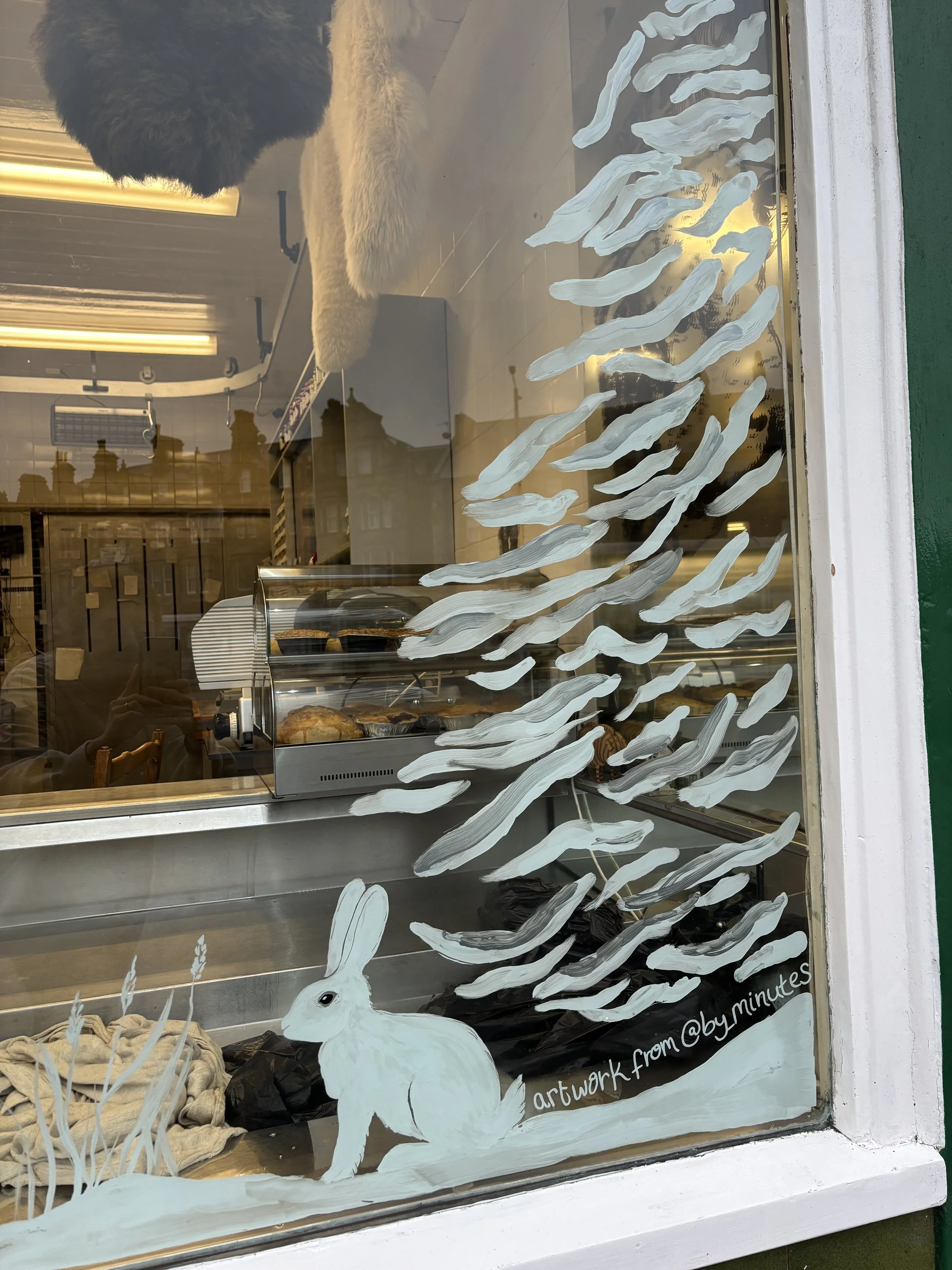 A window with an artistic painting of a white hare and white abstract lines representing trees, with the text 'artwork from @by_minutes'. Inside, bakery items can be seen behind the glass.
