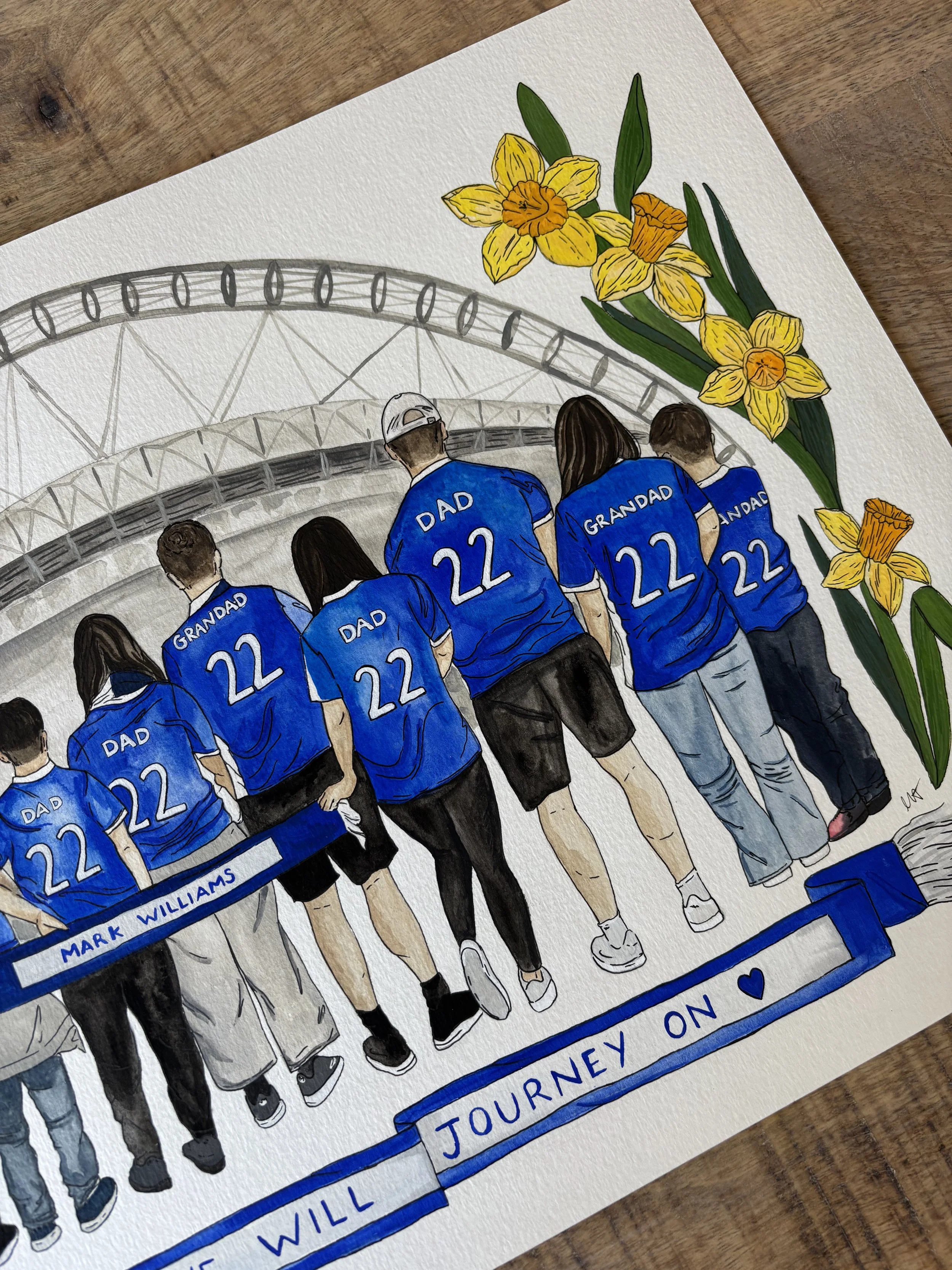 A drawing of a family, including children and an older man, all wearing matching blue shirts with the number 22 and the words 'Grandad' and 'Dad' on the back, walking together. The scene appears to be on a bridge, with a Ferris wheel in the background, and there are yellow daffodils on the side of the drawing. The text at the bottom reads 'We will journey on' with a heart symbol.